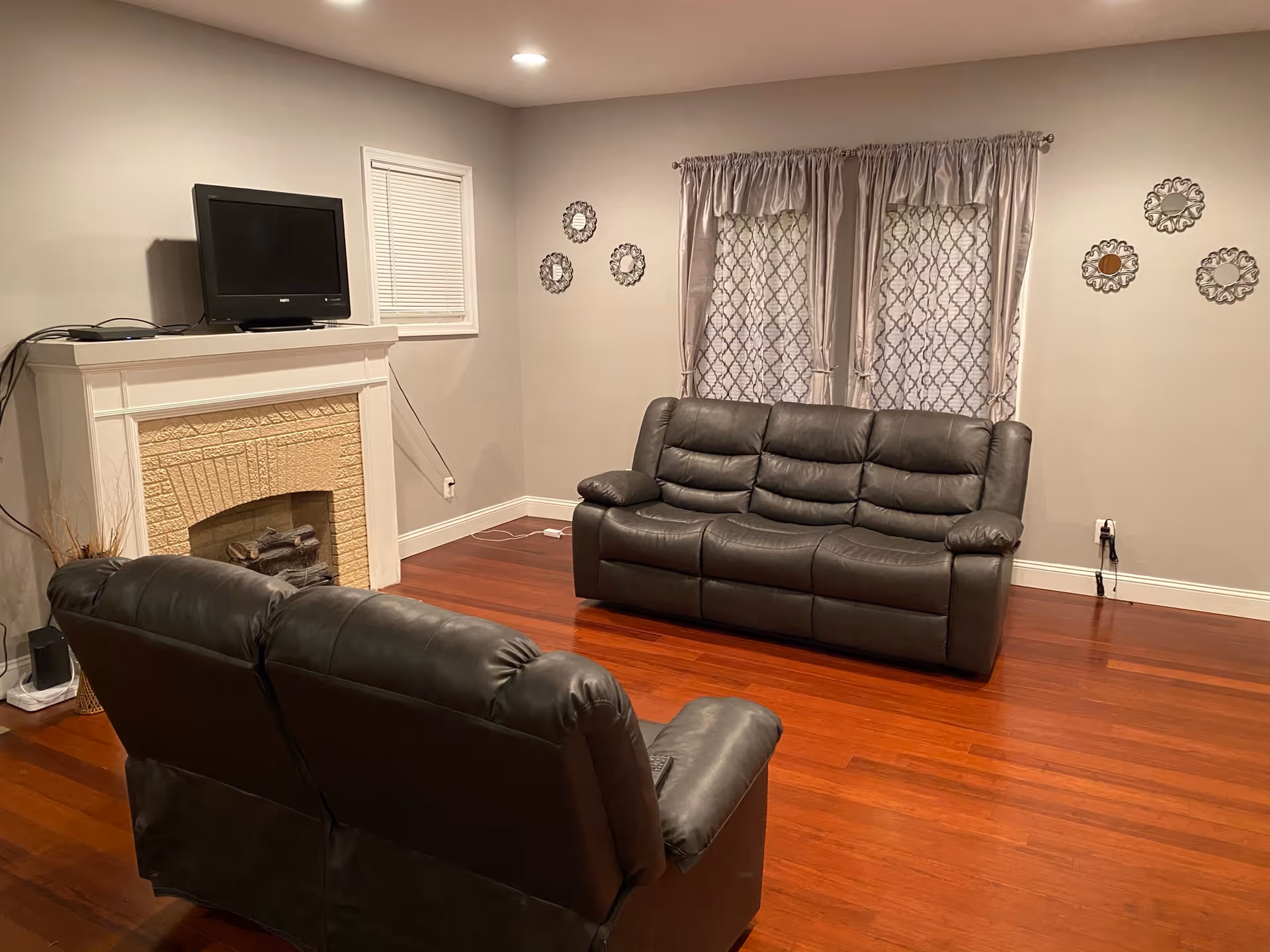 A living room with two dark leather sofas facing a white fireplace with a TV on top. The room has wooden floors, light gray walls, a small window with blinds, and two larger windows with patterned curtains. Decorative wall hangings are on the walls.