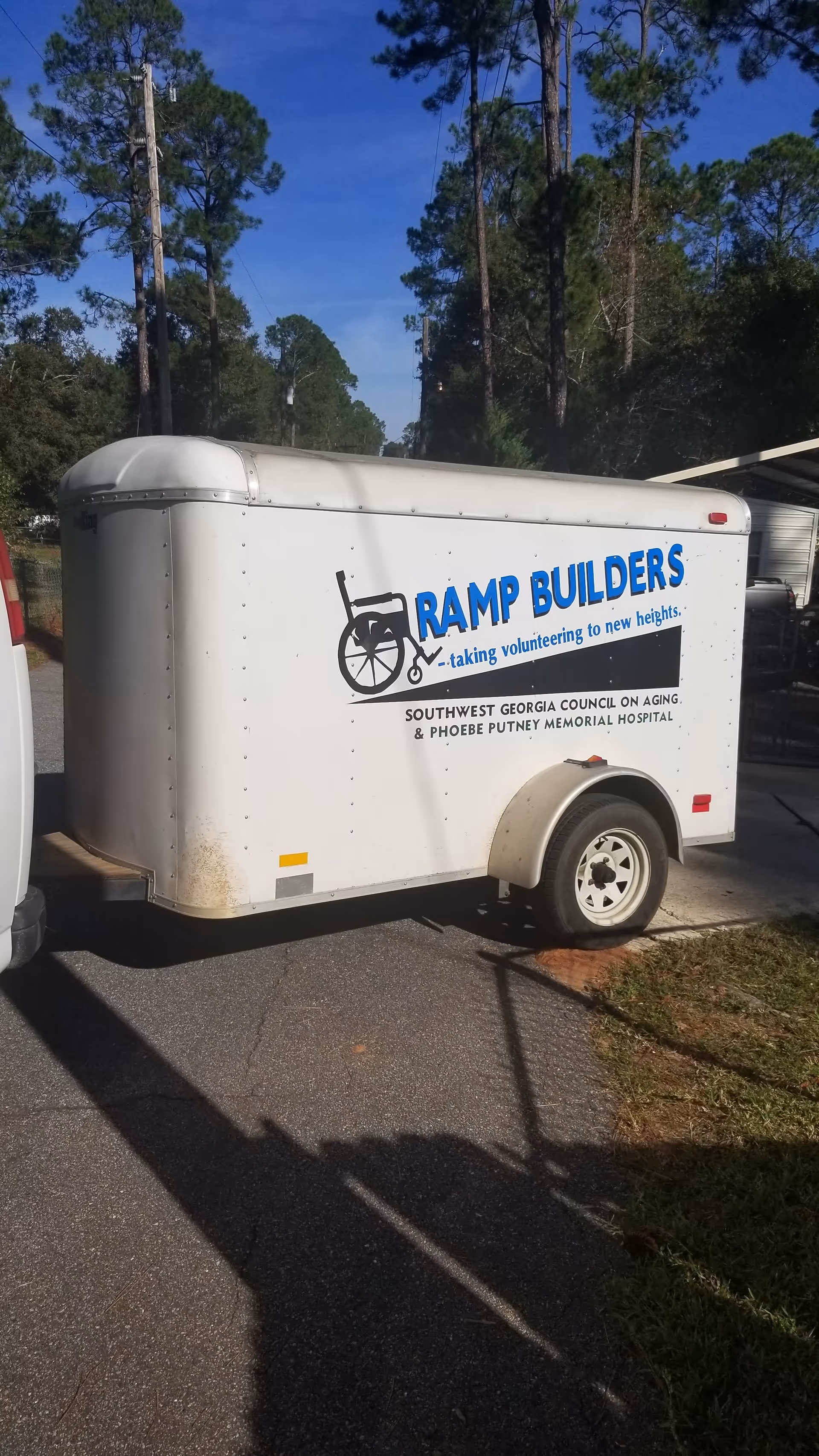 White trailer with Ramp Builders logo parked on a paved road amid tall pine trees.