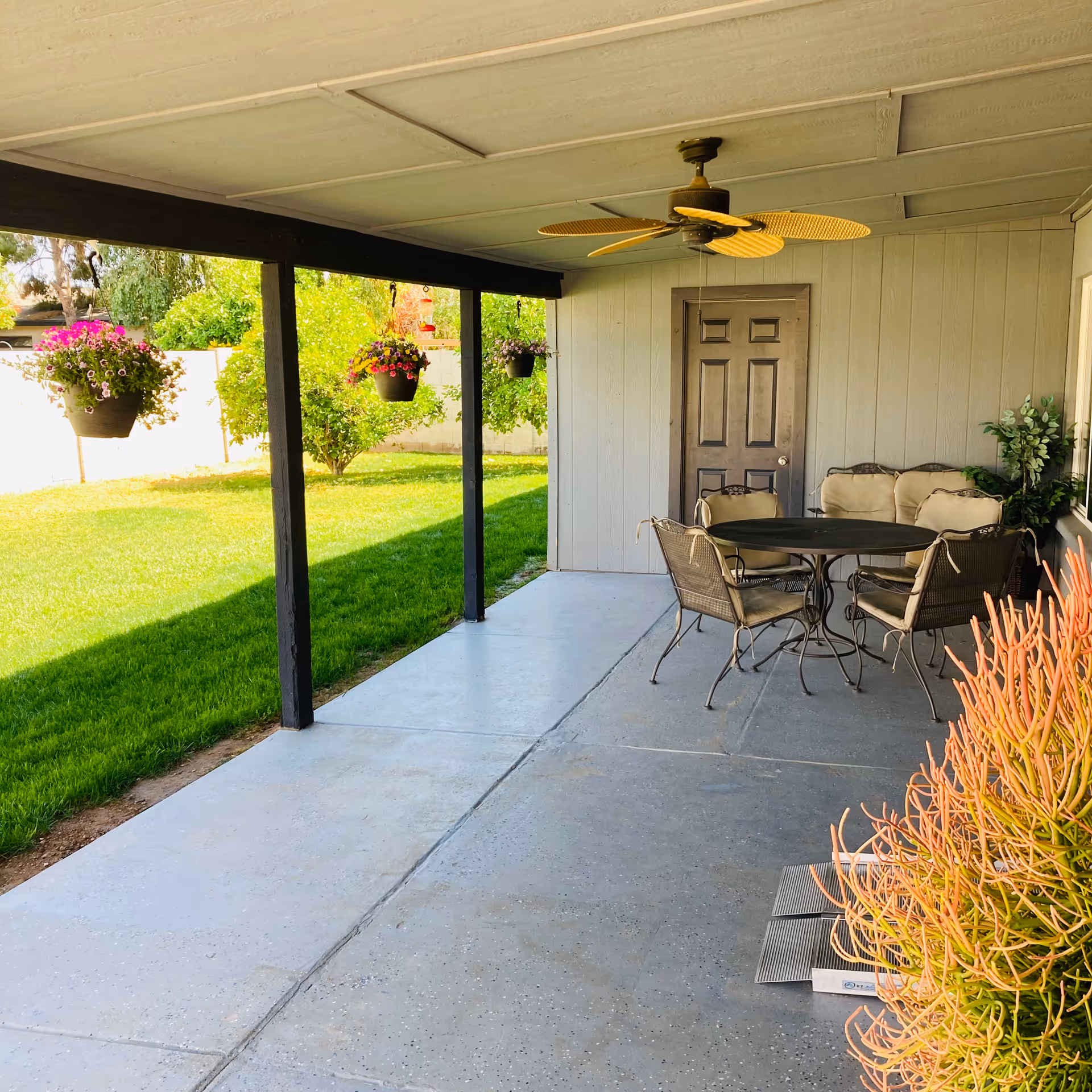 Covered patio with a round table, chairs, hanging plants, and a door on the wall.