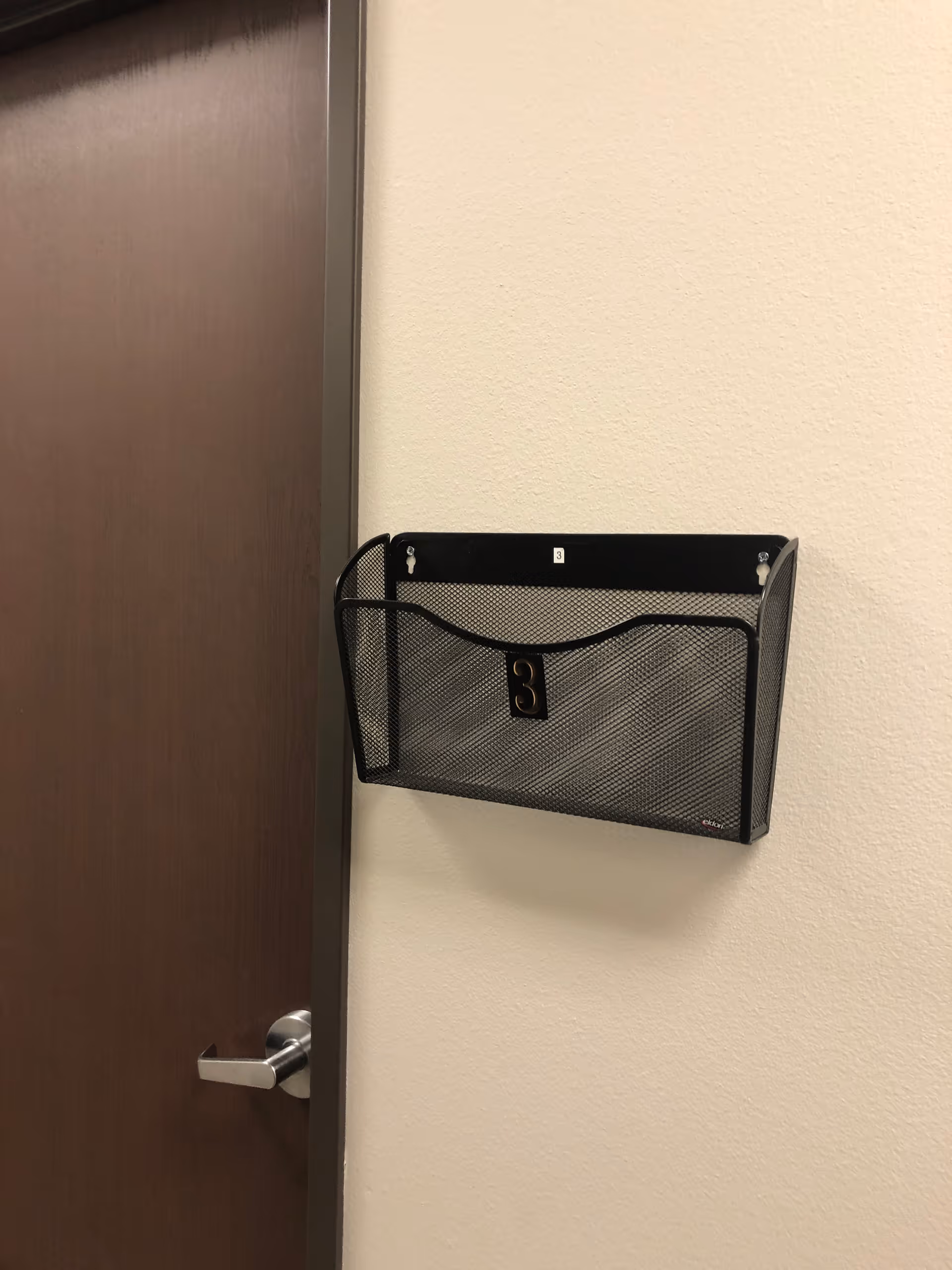 Metal mesh wall-mounted file holder labeled '3' beside a wooden door.