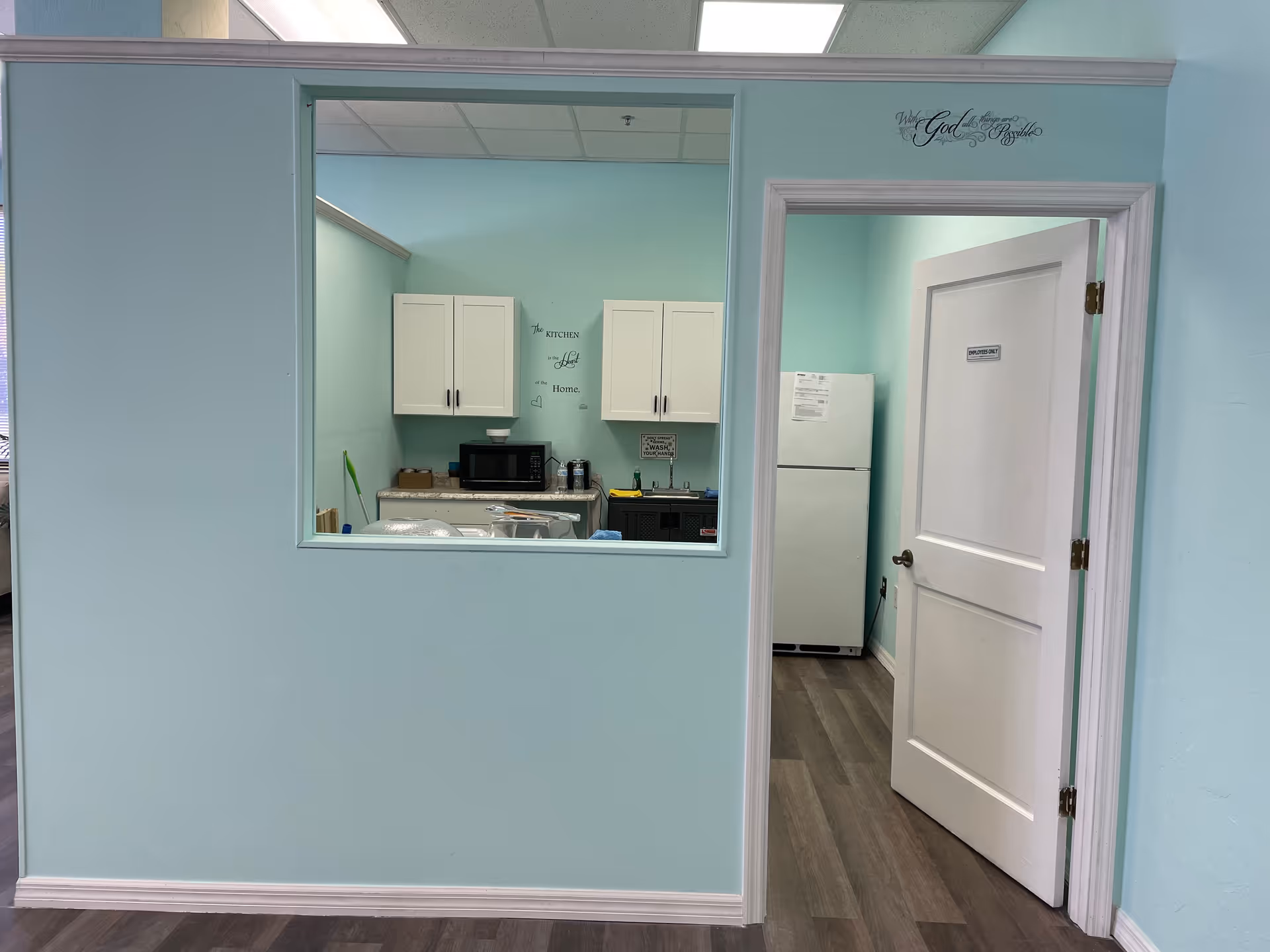 Bright turquoise wall frames a window into a small kitchen with cabinets and a fridge.