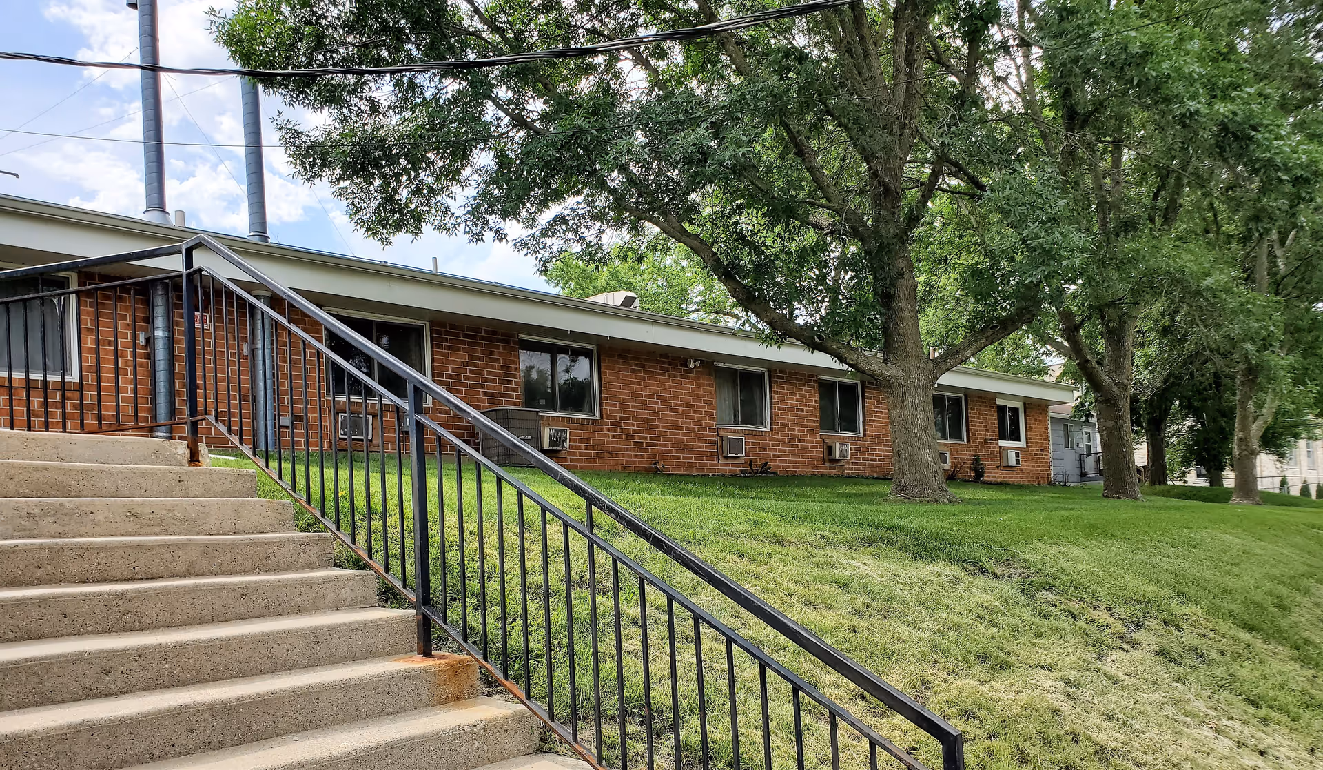 Front exterior of a single-story brick building with concrete steps and a black handrail, grassy lawn, and large trees.