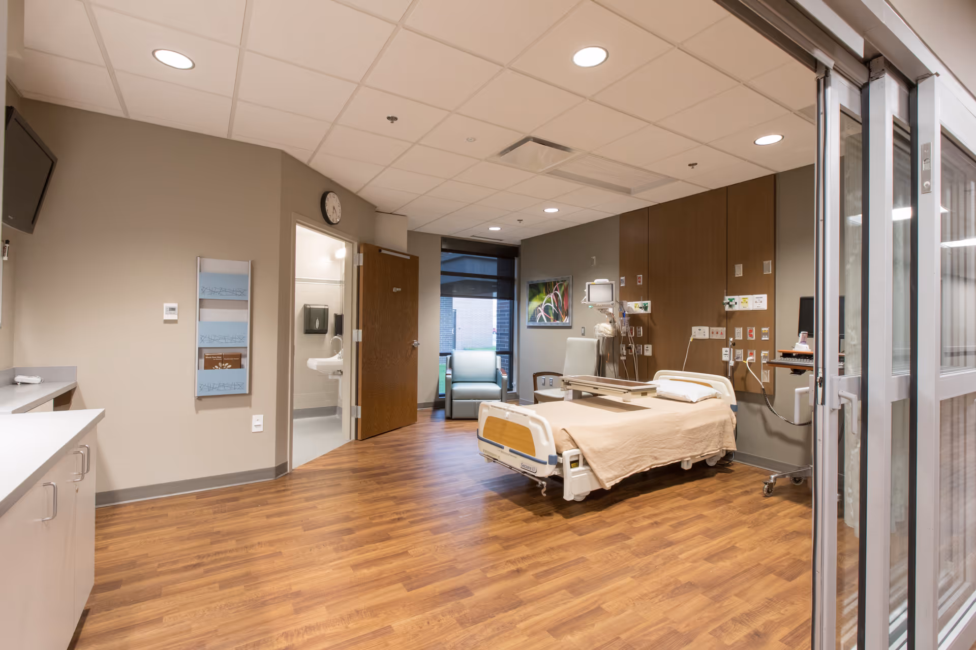 Hospital patient room with adjustable bed, medical equipment, wood flooring, and glass door.