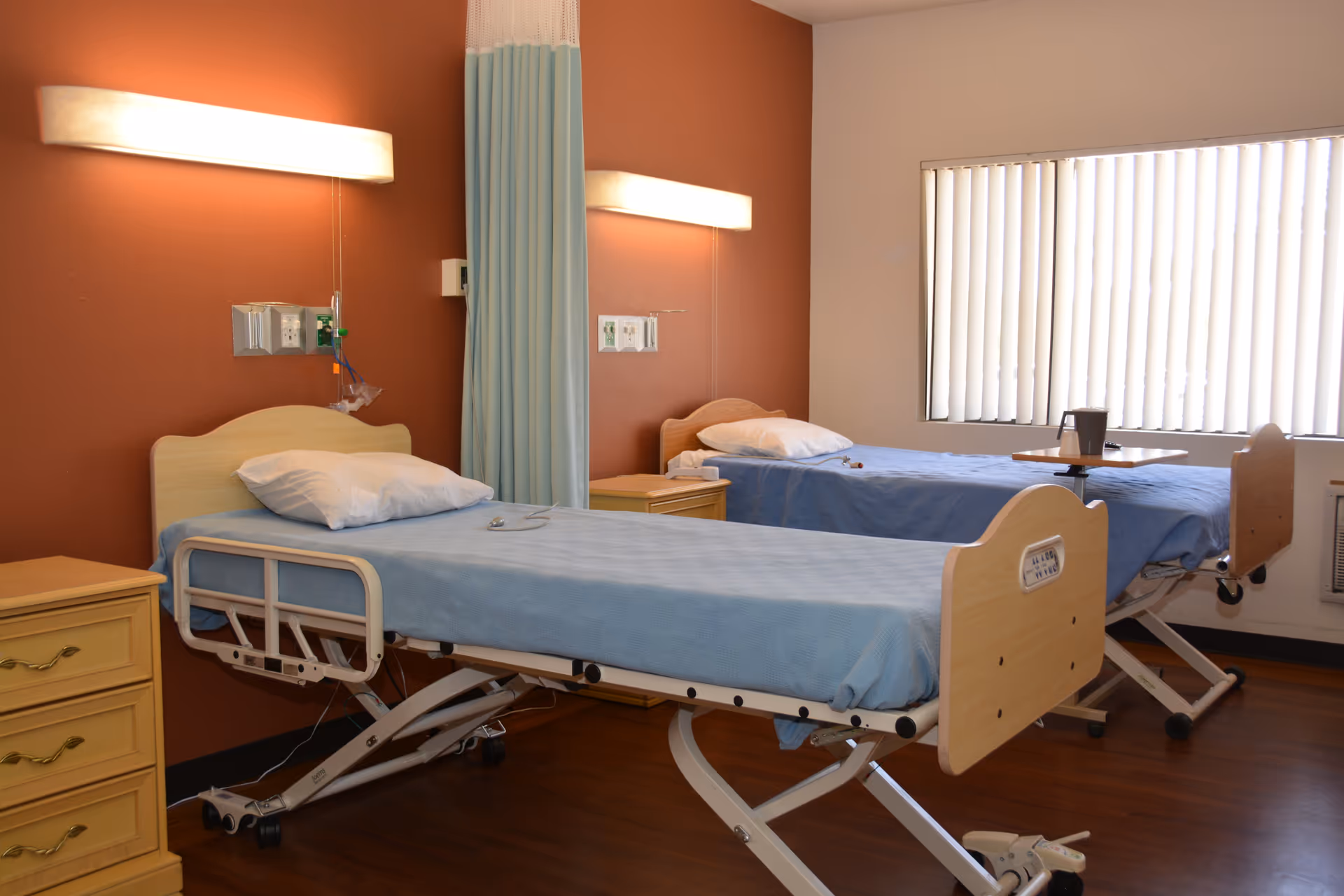 Two hospital beds with blue sheets in a bright patient room with privacy curtains.