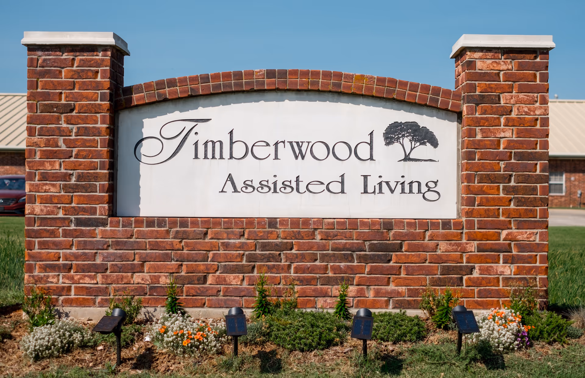A brick sign for Timberwood Assisted Living stands between two brick pillars with landscaping.