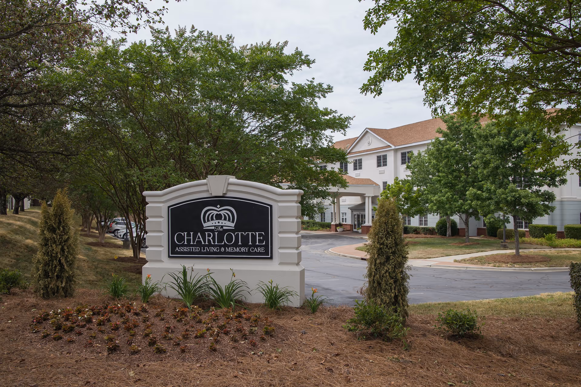 Sign reading 'The Charlotte Assisted Living & Memory Care' at the front of the facility with the building and driveway behind it.