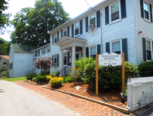 Exterior view of Wadleigh House Assisted Living, a two-story white building with black shutters and a small porch supported by columns. There are bushes and flowers planted along the front, a brick walkway, and a sign displaying the facility's name and phone number.