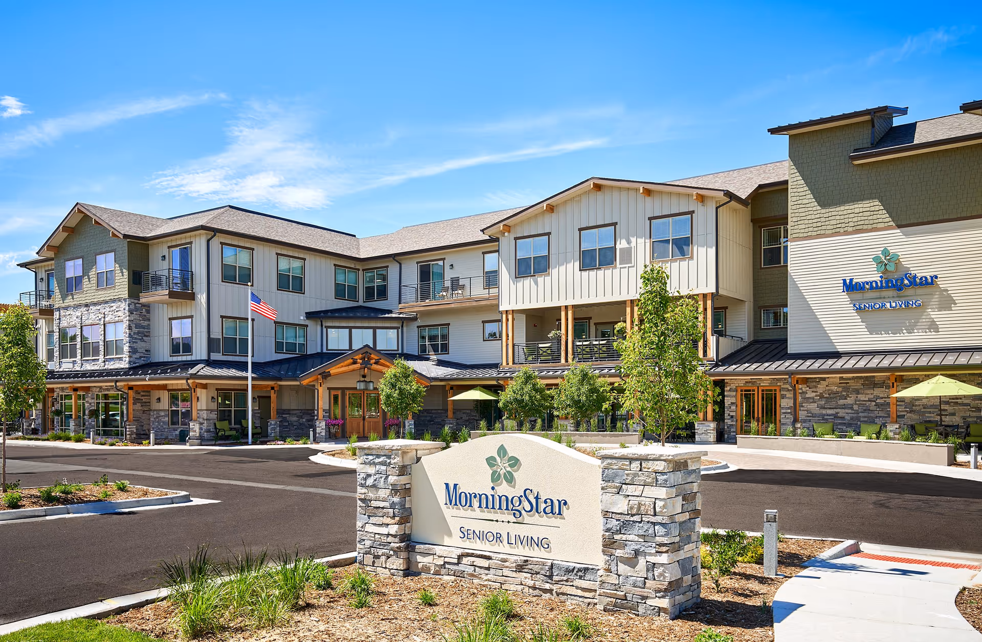 Exterior view of MorningStar Senior Living of Arvada, a modern three-story senior living facility with a stone and siding facade, balconies, an American flag, landscaped grounds, and a clear blue sky.