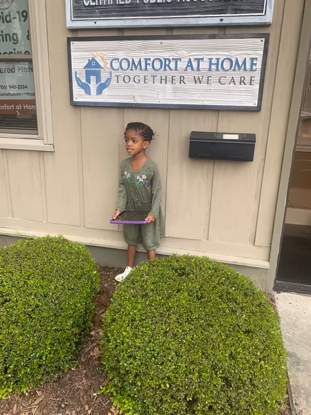 Young girl stands outside a Comfort At Home sign on the building front.
