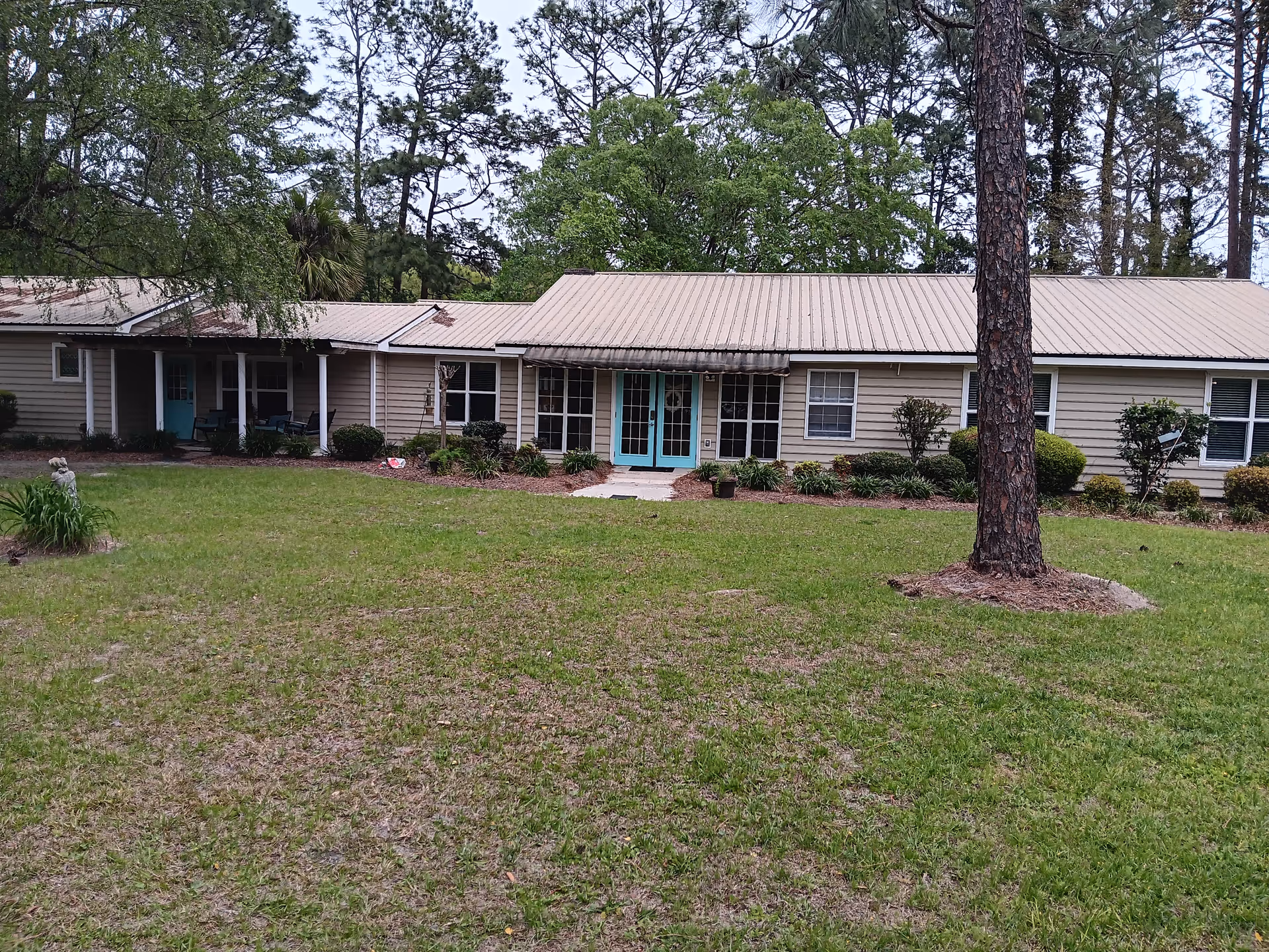 Single story senior living facility front with turquoise doors, manicured lawn, and tall trees.