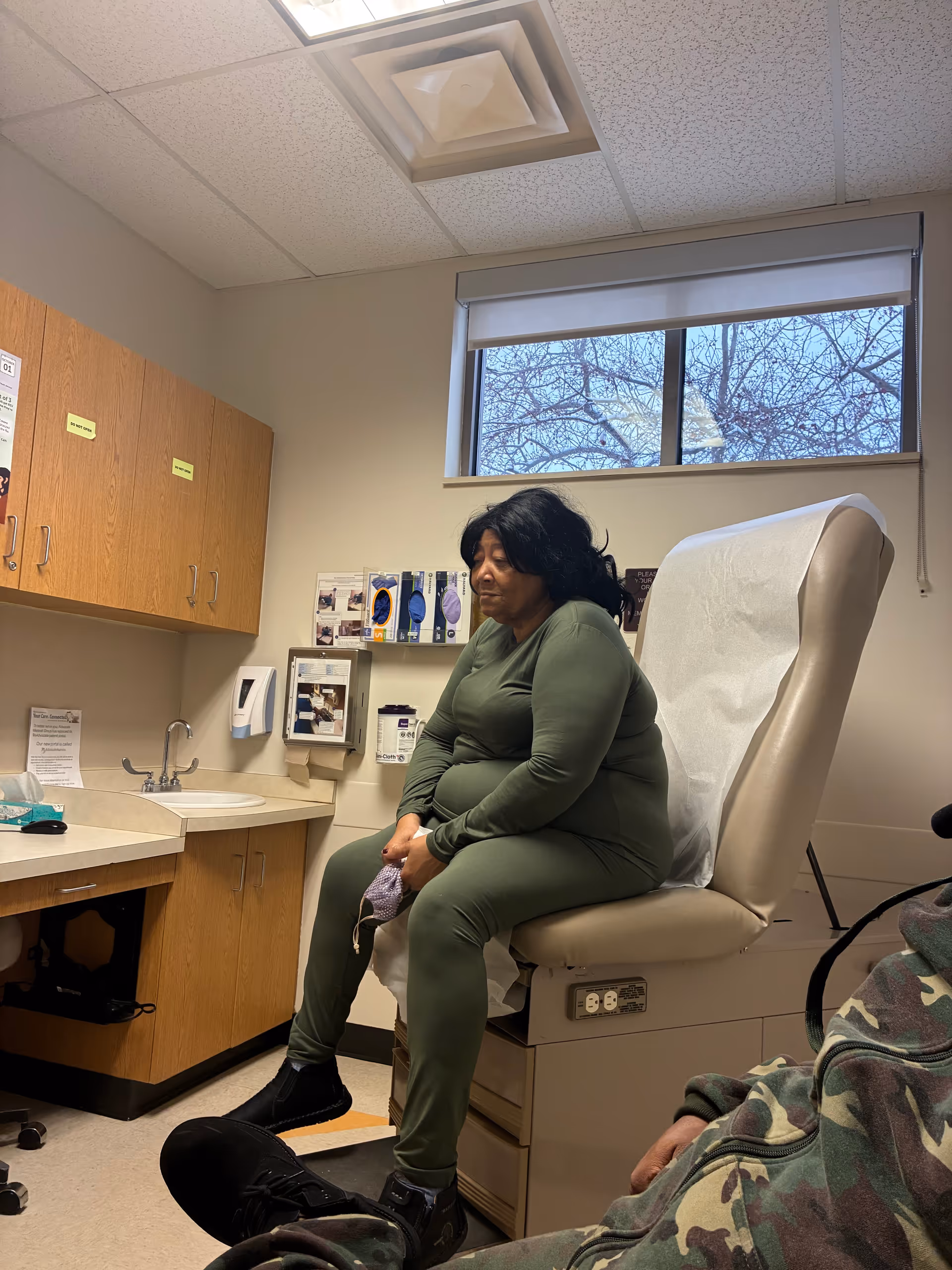 A woman sits on an exam chair in a clinic room with cabinets and a window.