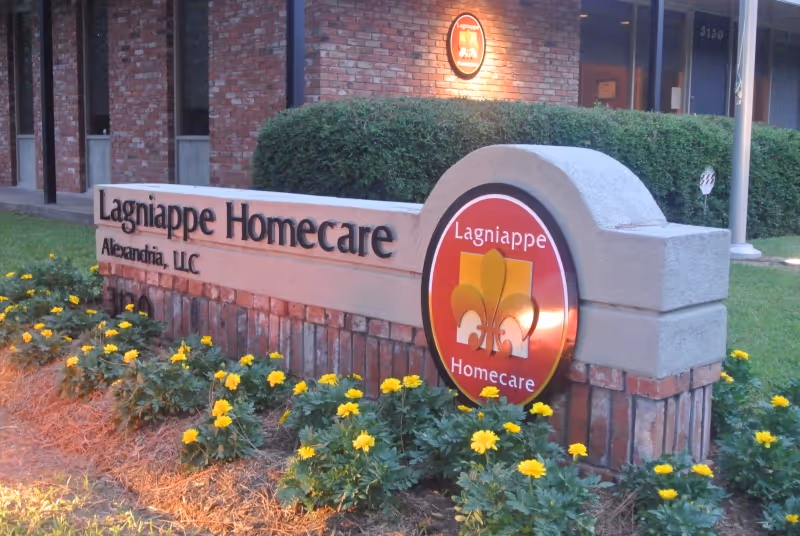 Stone sign reading Lagniappe Homecare with yellow flowers and brick building facade behind.