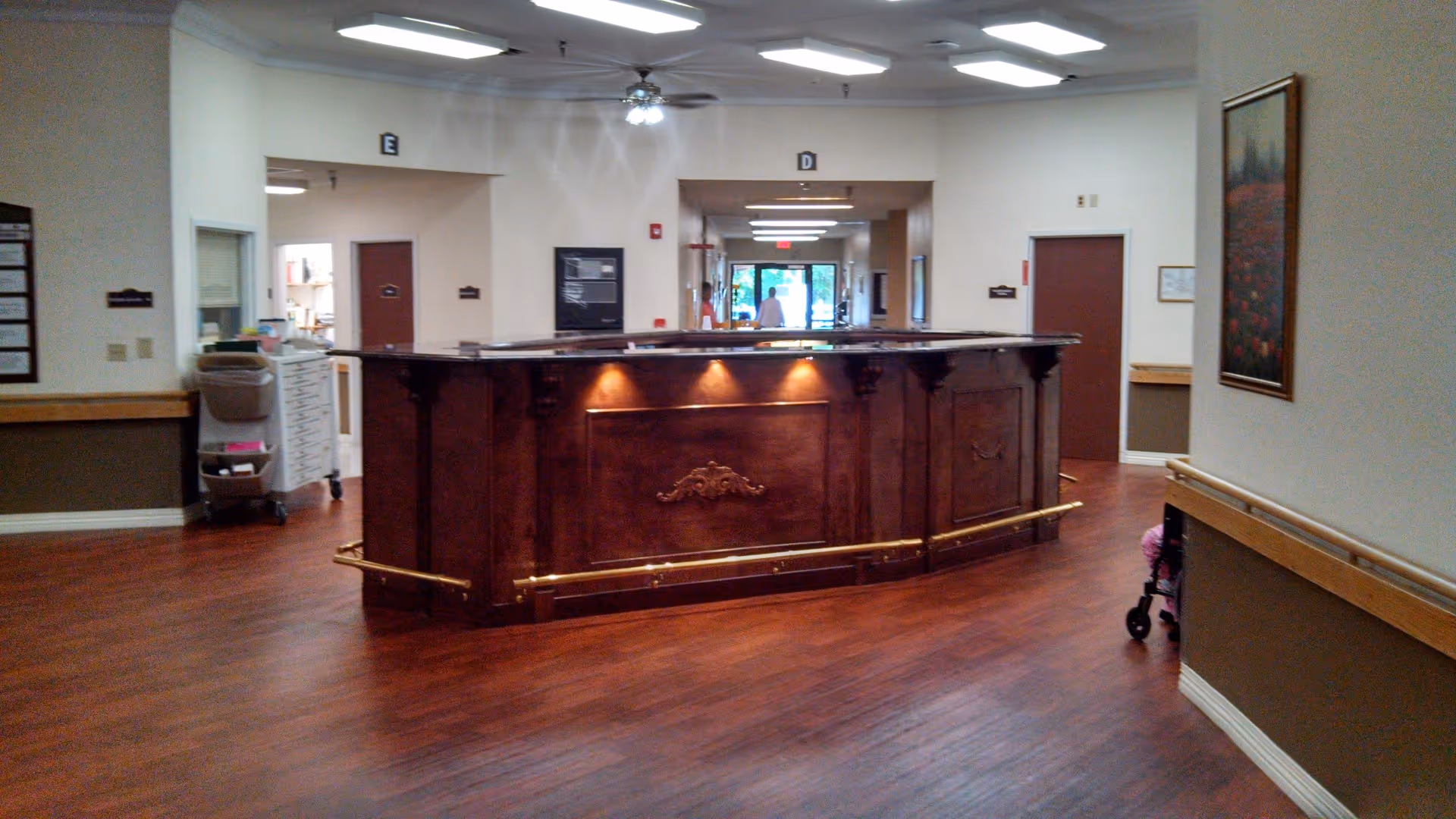 Interior view of a healthcare facility reception area with a wooden reception desk in the center, wood flooring, beige walls, ceiling lights, and doors labeled with letters E and D. A person is visible in the background near the exit door.