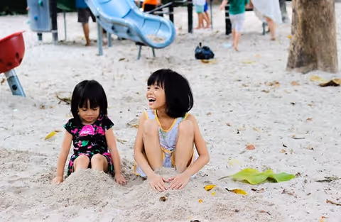 Two young girls sit in sand at a playground, smiling near a slide.