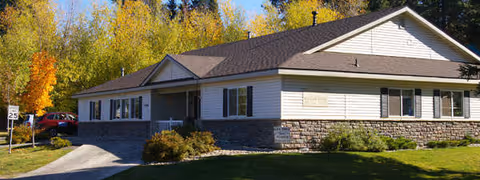Single-story light-colored cottage-style building with a stone veneer base, front porch, driveway and trees in the background.