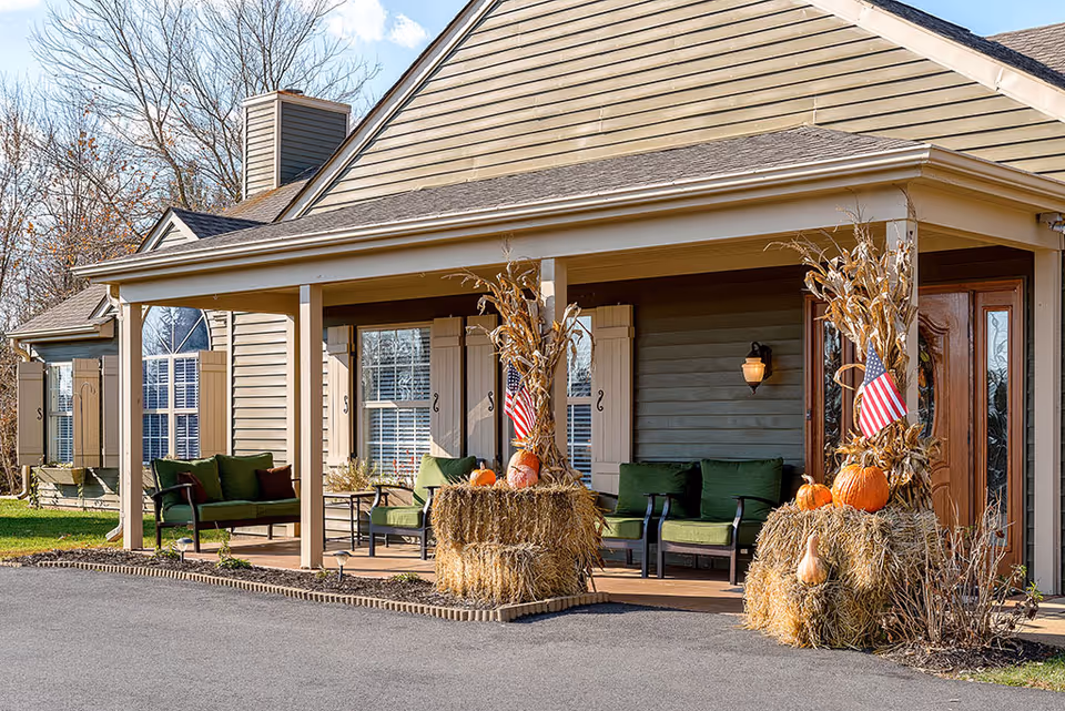 Front porch of a senior living facility decorated with hay bales, pumpkins, dried corn stalks, and American flags. The porch has green cushioned chairs and a wooden door, with beige siding and white window shutters on the building.