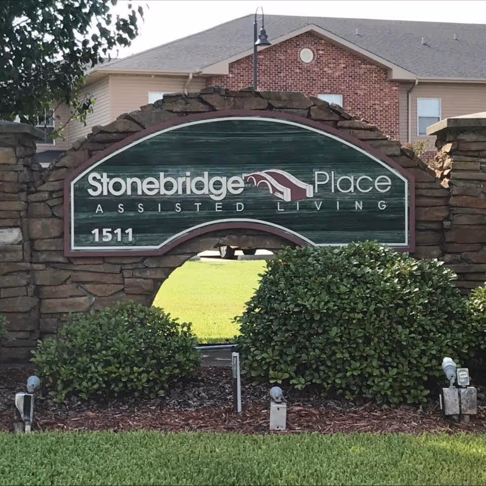 Stonebridge Place Assisted Living facility entrance sign made of stone and wood with bushes and grass in front, and a residential building in the background.