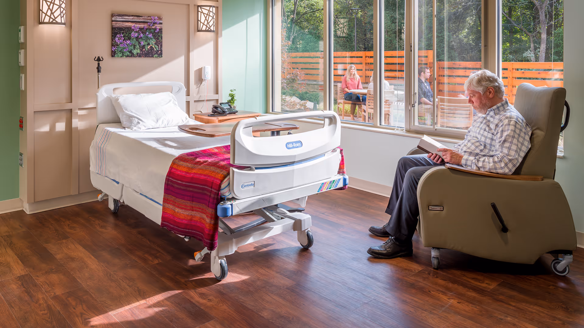 Hospice patient room with a hospital bed, a man reading in a recliner, and large windows.
