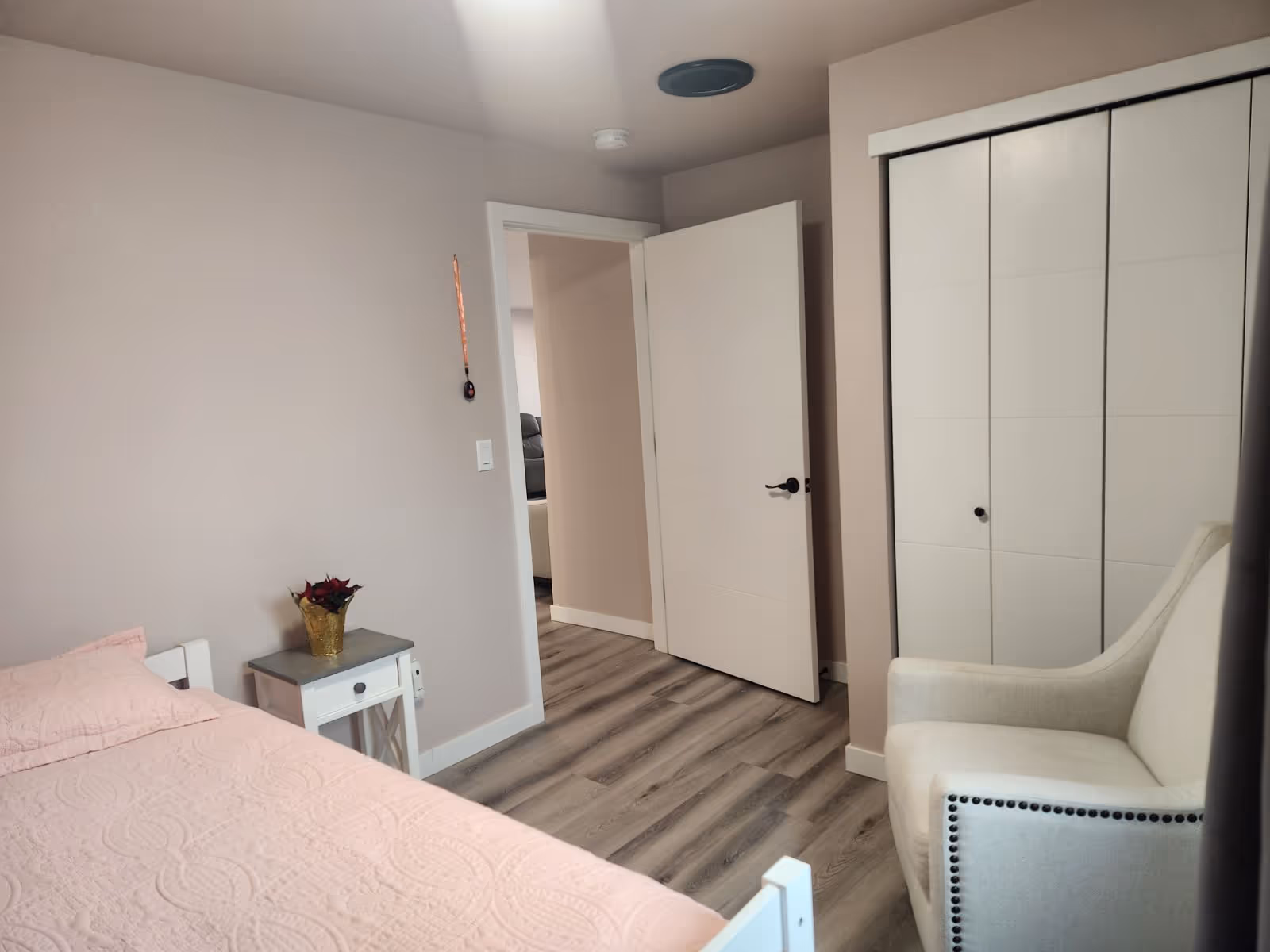 A cozy bedroom featuring a pink bed, a small side table, and an armchair, with an open door leading to another room.