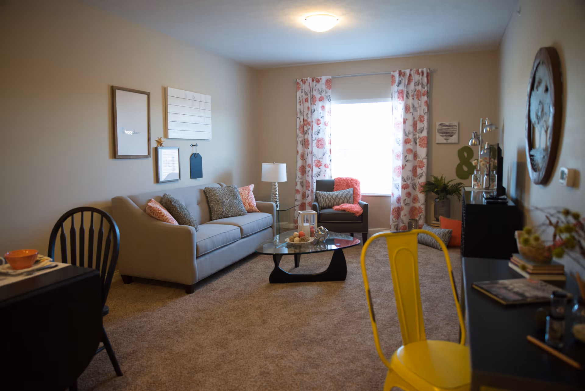 A cozy living room with a beige sofa adorned with patterned cushions, a glass coffee table with decorative items, a gray armchair with a coral throw, floral curtains on the window, a floor lamp, wall art, and a yellow chair in the foreground. The room has beige walls and carpeted flooring.
