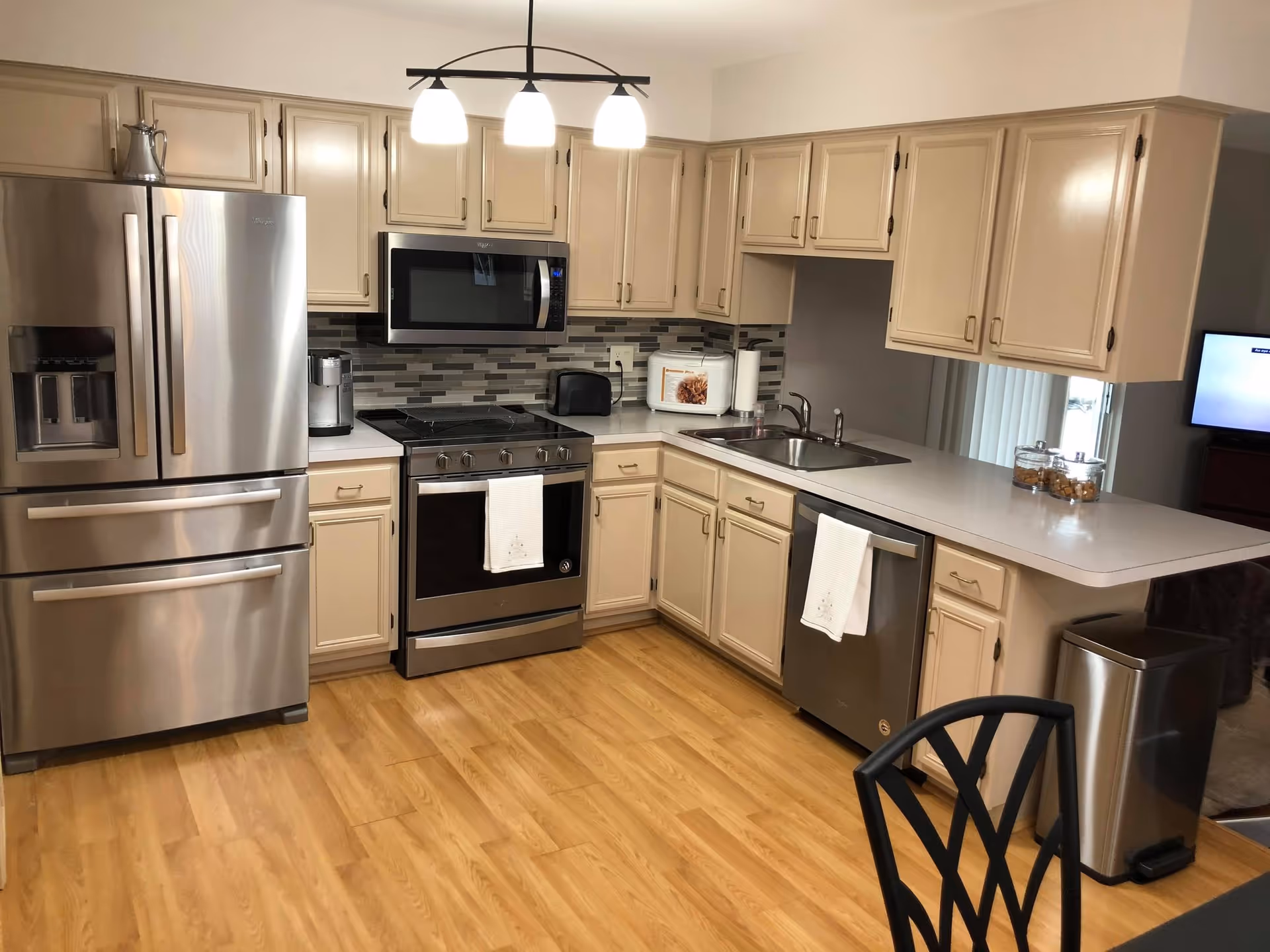 A modern kitchen with light beige cabinets, stainless steel refrigerator, stove, microwave, and dishwasher. The kitchen has a wooden floor, a double sink, a countertop with a paper towel holder, and a few jars with snacks. There is a black chair partially visible in the foreground and a trash can near the counter.