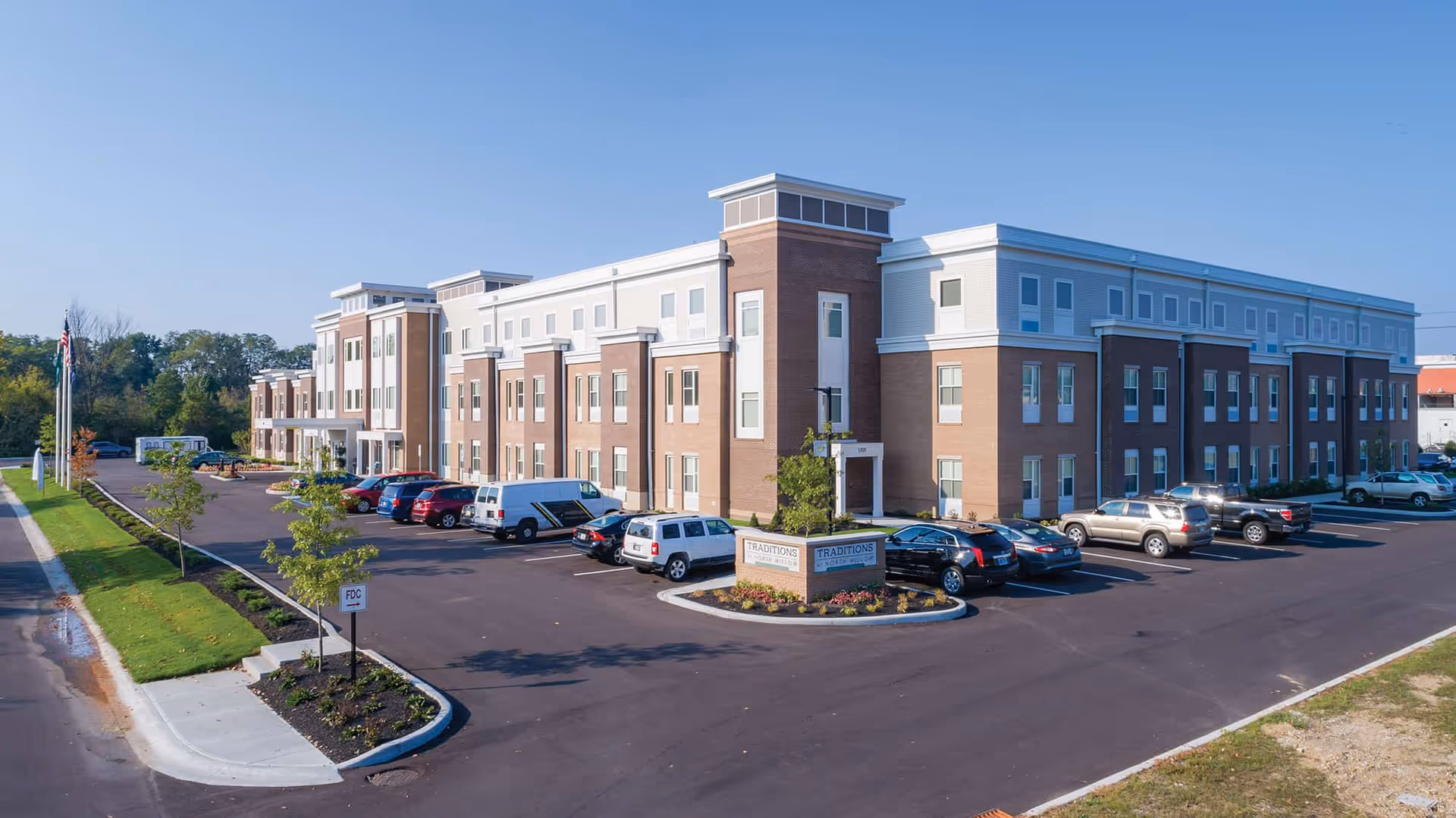 Three-story brick-and-white senior living building with a parking lot and landscaped entrance under a clear blue sky.