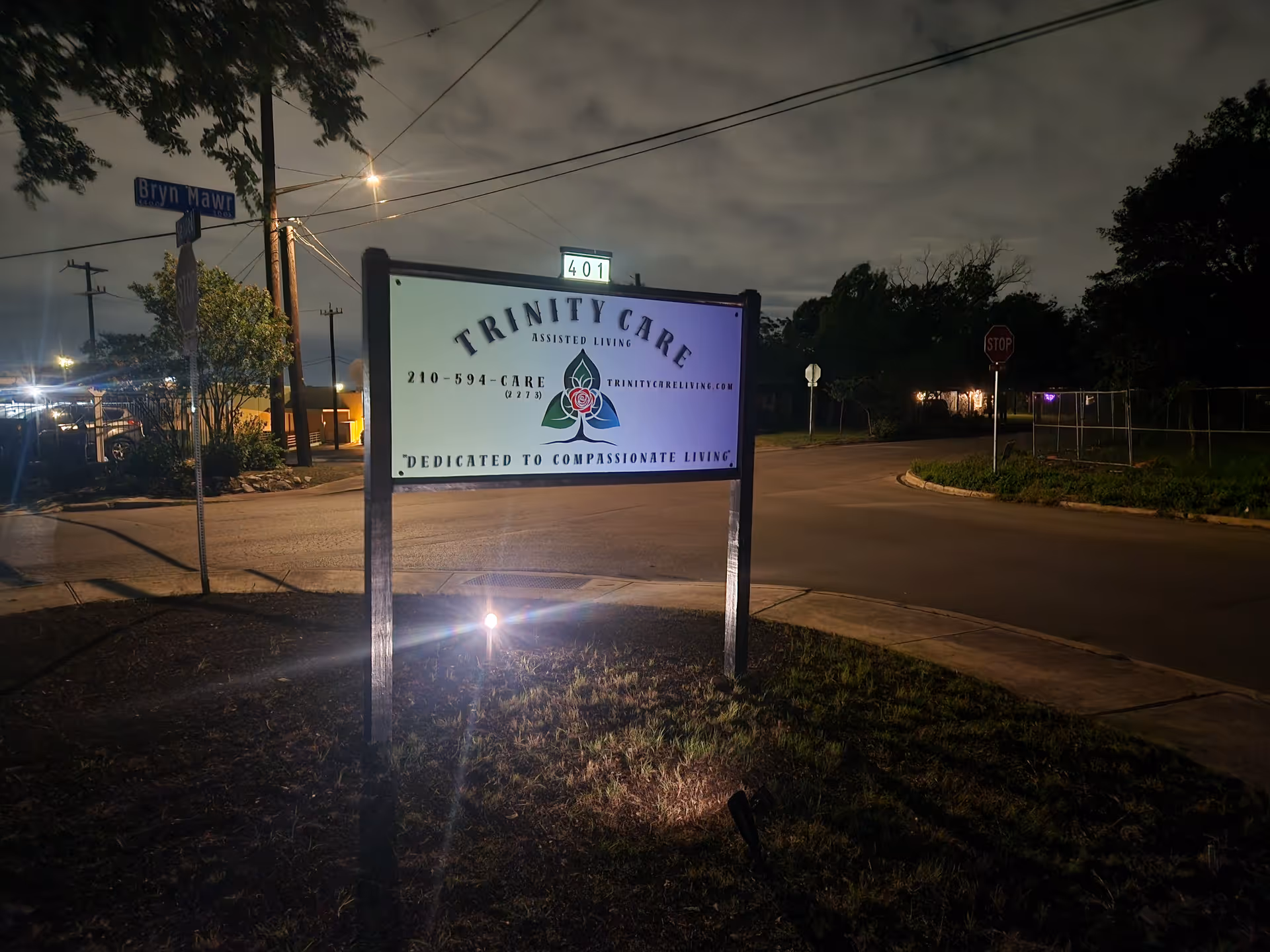 Nighttime exterior street scene with an illuminated Trinity Care sign on a street corner.
