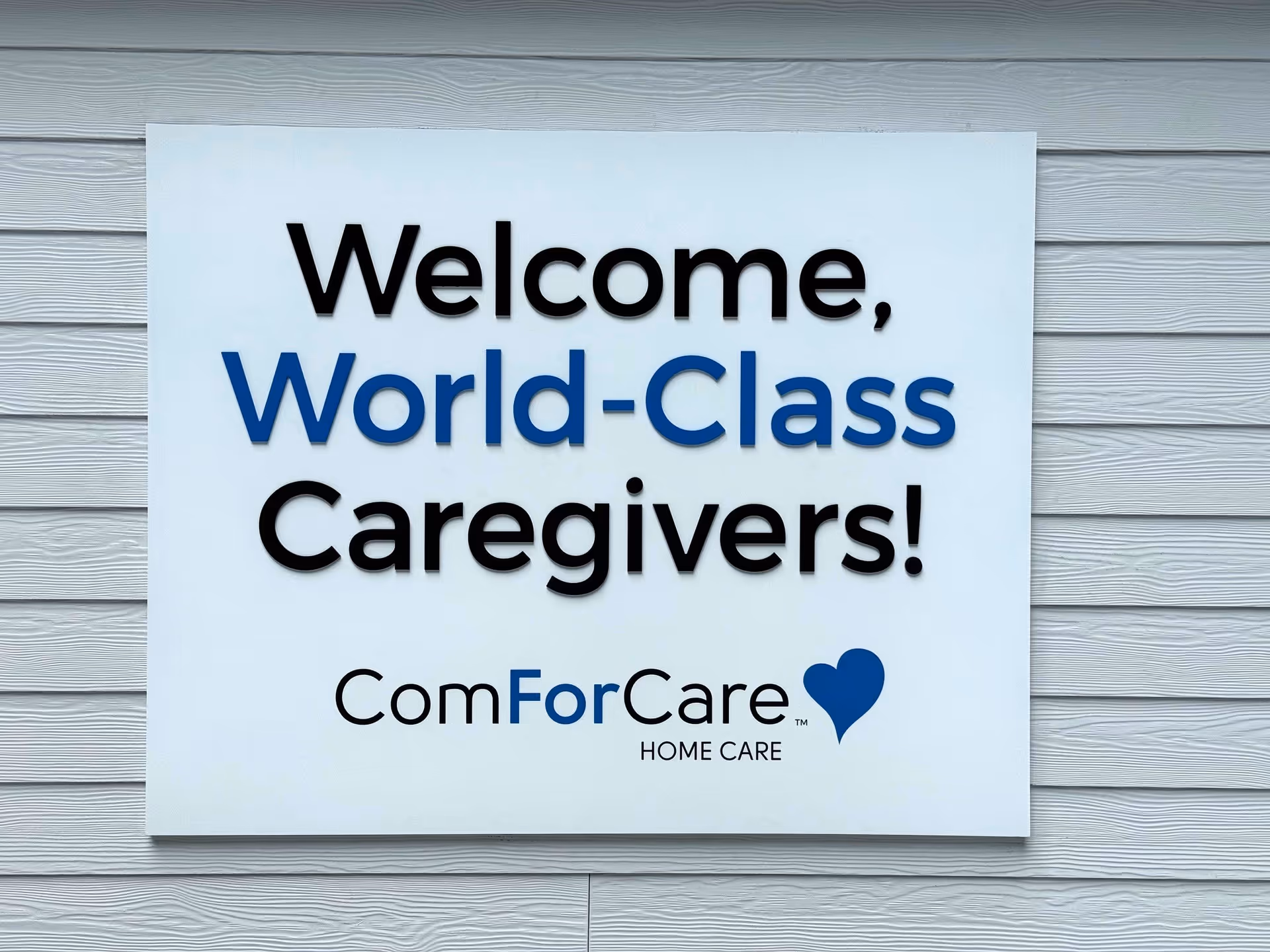 White square sign on light-gray wooden wall reading Welcome, World-Class Caregivers, ComForCare.