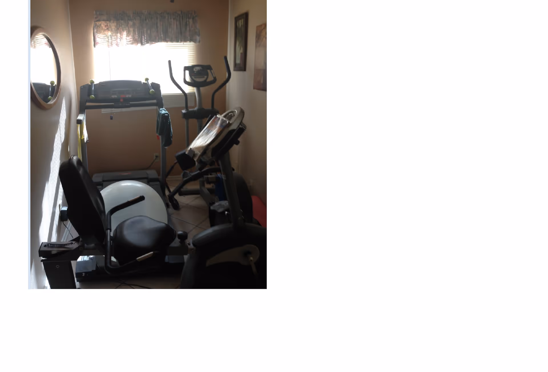 Small exercise room with a recumbent stationary bike, treadmill, elliptical machine, and an exercise ball. A window with a floral valance lets in natural light, and a round mirror hangs on the left wall.