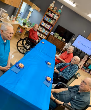 Five elderly men sitting around a long table covered with a blue tablecloth in a common room. Each man has a plate with a donut in front of him. The room has a bookshelf filled with books and decorative birdhouses, a television displaying the Novartis logo, and various other furniture and decorations.