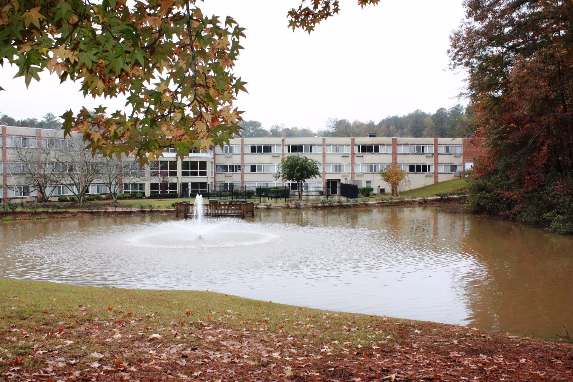 View of a large pond with a water fountain in the center, surrounded by grass and autumn leaves. In the background, there is a multi-story building with many windows, partially obscured by trees with fall foliage.