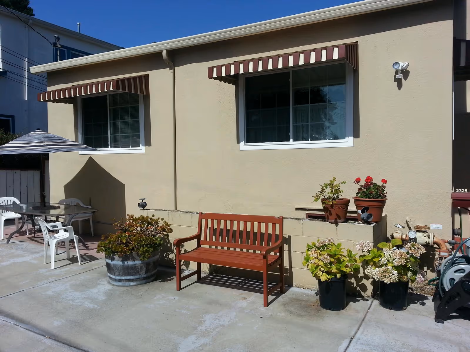 Outdoor patio with a wooden bench, plastic chairs, and potted plants beside a beige building.