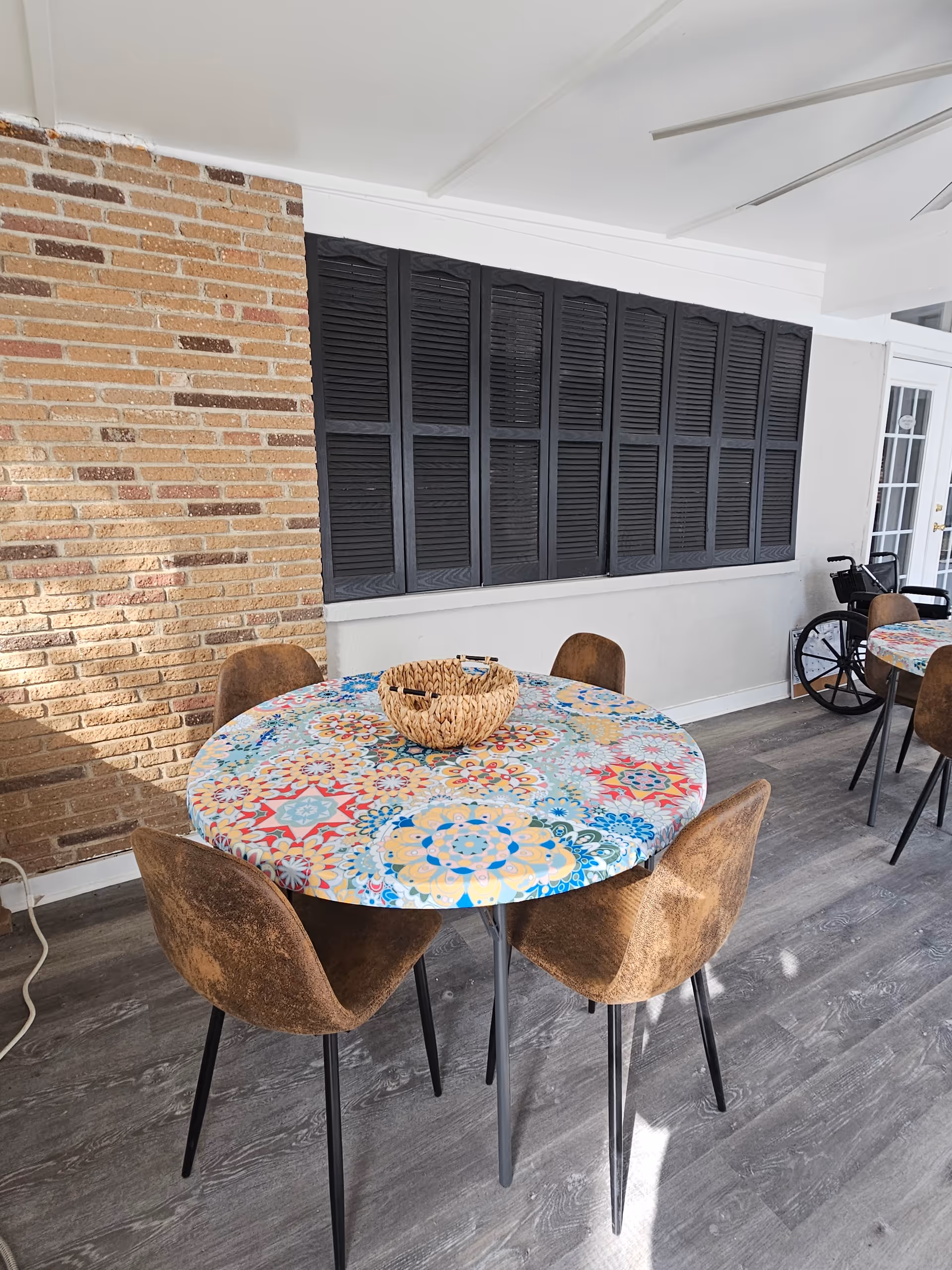 Outdoor seating area with round patterned table, wicker chairs, brick wall, and a wheelchair nearby.