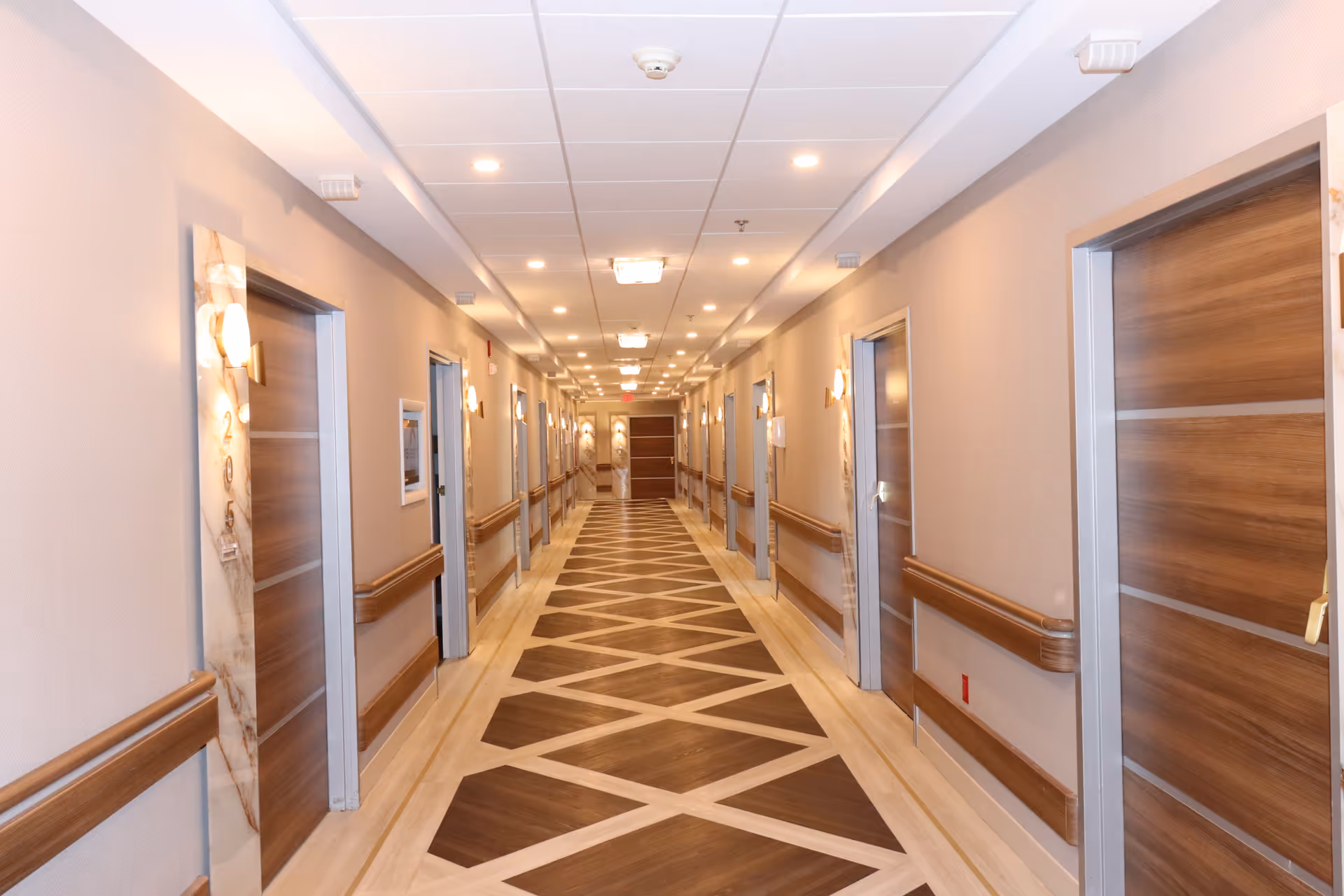 Long beige hallway in a senior living facility with doors, wood railings, and patterned floor.