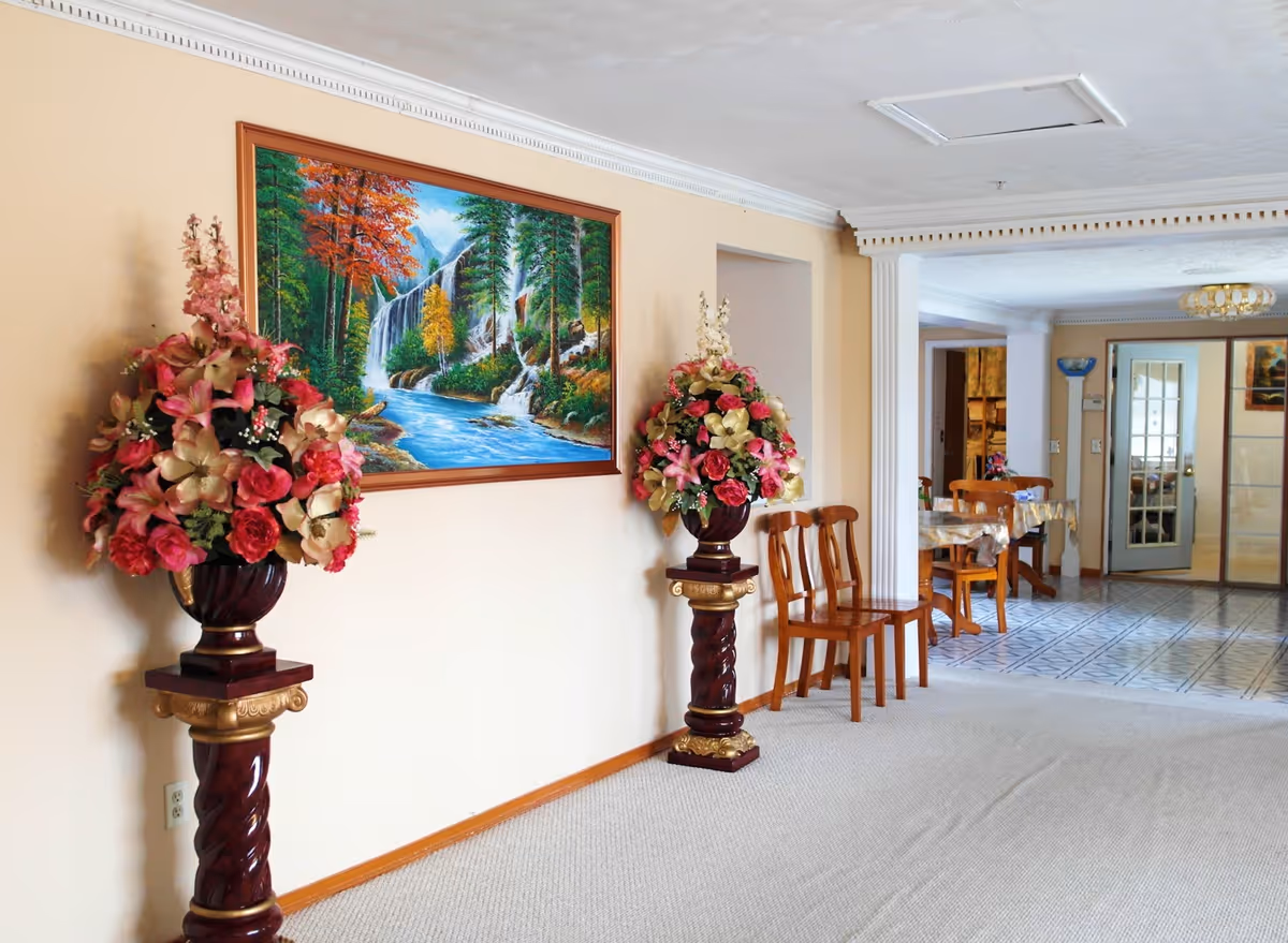 Interior hallway with tall flower arrangements, a landscape painting, and a dining area in the background.