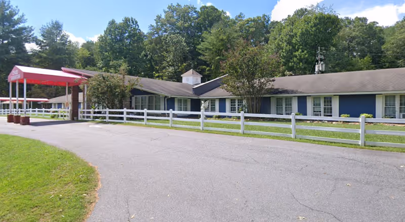 A single-story blue assisted living building with a white fence and red entrance canopy.