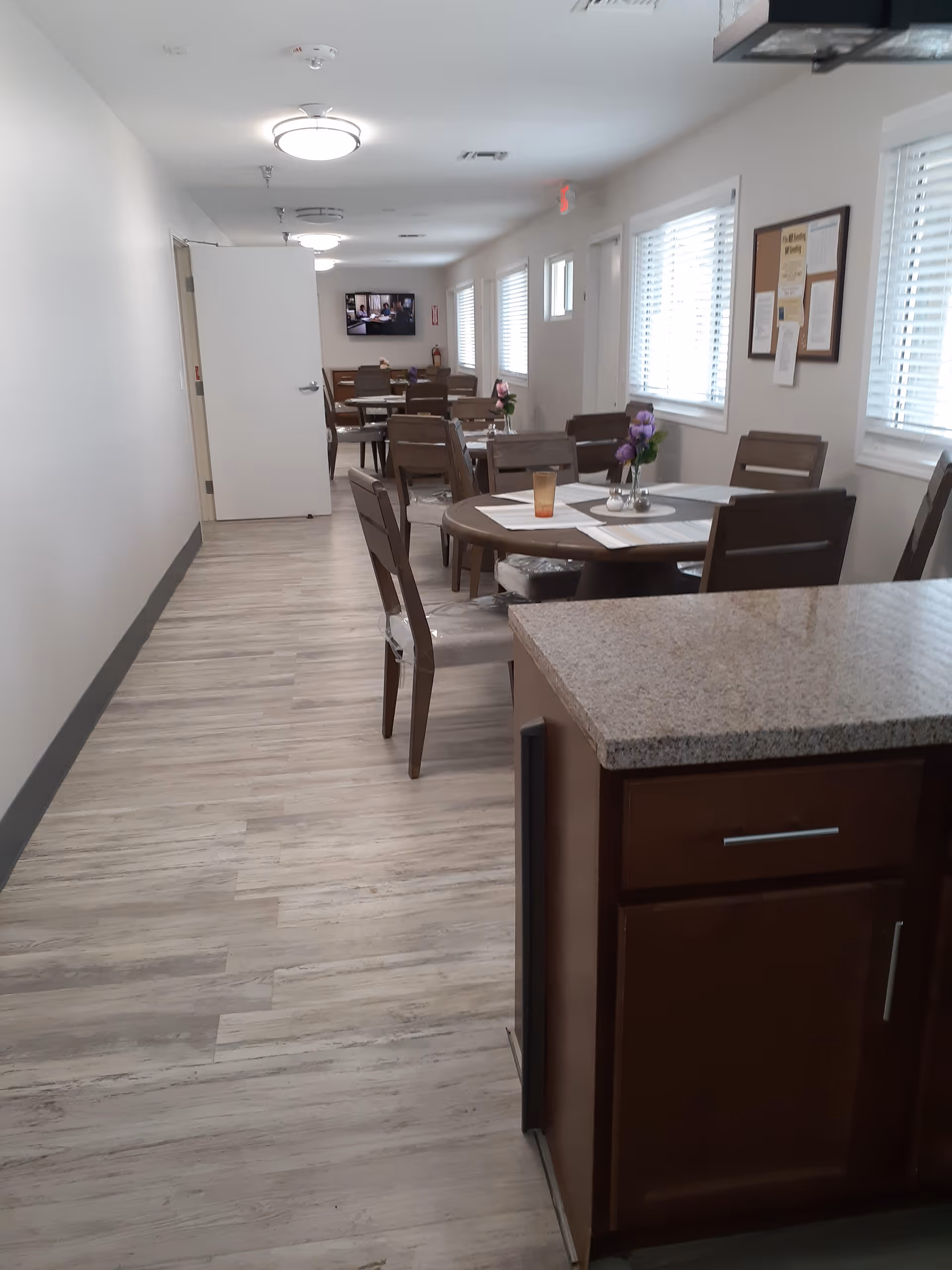 Interior view of a dining area in a senior living facility with several wooden tables and chairs arranged along a hallway. The tables have placemats and small flower vases. There are windows with blinds on the right side and a television mounted on the far wall. The floor is light-colored wood, and there is a kitchen counter with a granite countertop in the foreground on the right.