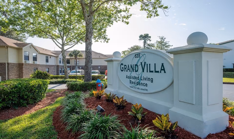Outdoor view of the entrance sign for Grand Villa Assisted Living Residence surrounded by landscaped greenery, with a two-story residential building and trees in the background under a clear sky.