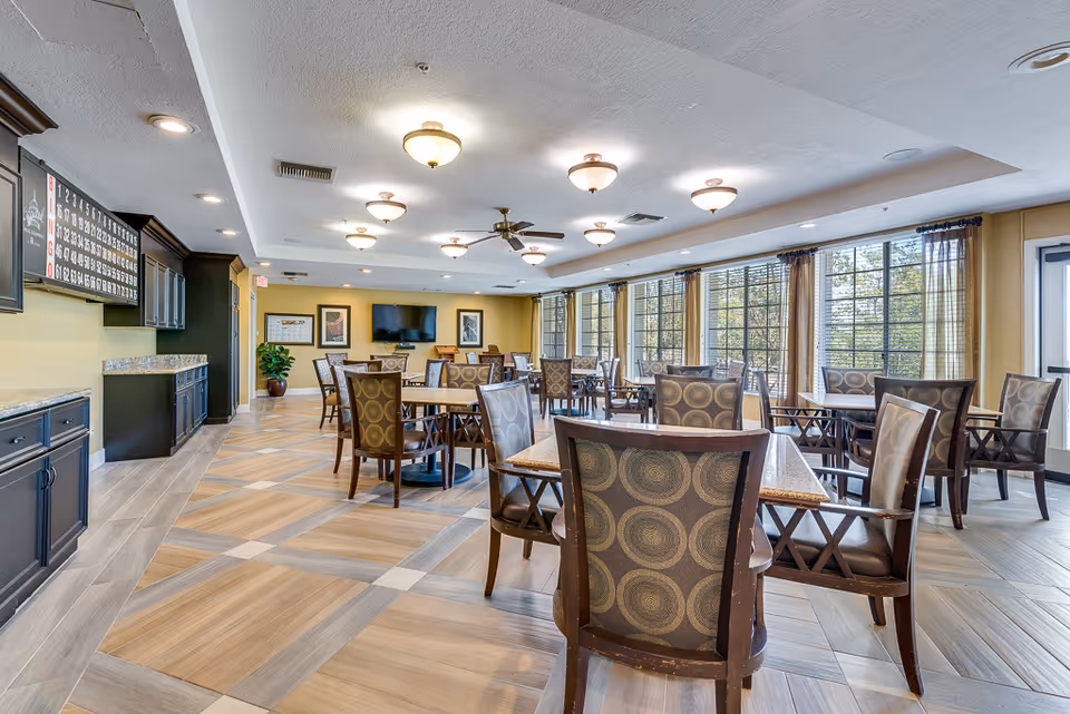 A spacious dining room in a senior living facility with multiple tables and chairs arranged neatly. The room has large windows with sheer curtains allowing natural light to fill the space. The ceiling features several light fixtures and a ceiling fan. There is a large bingo board on one wall and a flat-screen TV mounted on another wall. The floor has a patterned tile design, and there are cabinets along one side of the room.