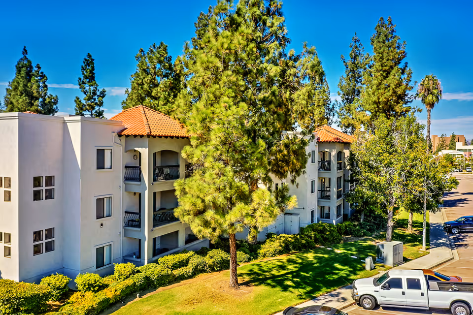 Three-story Mediterranean-style building with red tile roofs, balconies, trees, and parked cars in front.