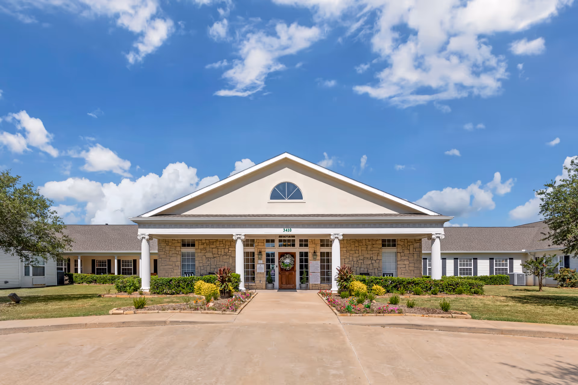 Front entrance of a single-story senior living building with white columns, a stone facade, landscaped flower beds, and a circular driveway under a blue sky.