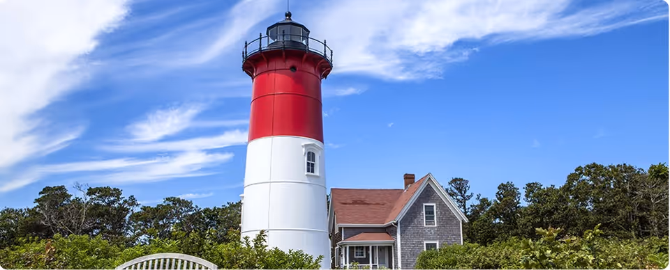 Red and white lighthouse beside a grey house under blue sky with green shrubs.