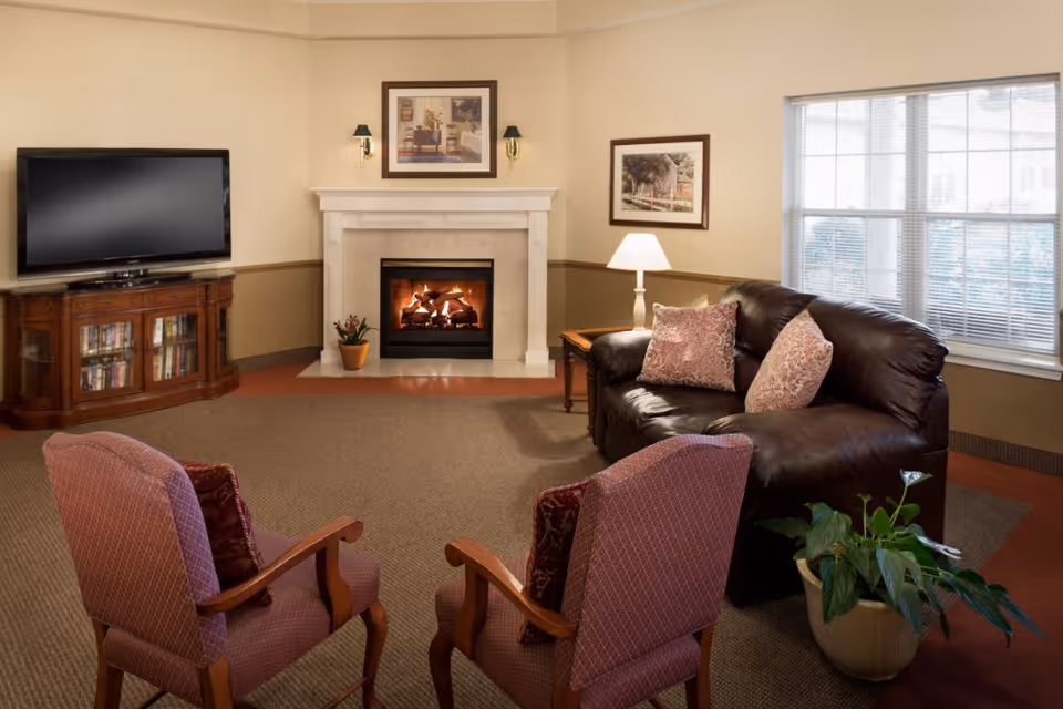 A cozy living room with a lit fireplace, a dark brown leather sofa with two patterned pillows, two upholstered armchairs, a wooden TV stand with a flat-screen television, a side table with a lamp, framed artwork on the walls, a large window with blinds, and a potted plant on the floor.