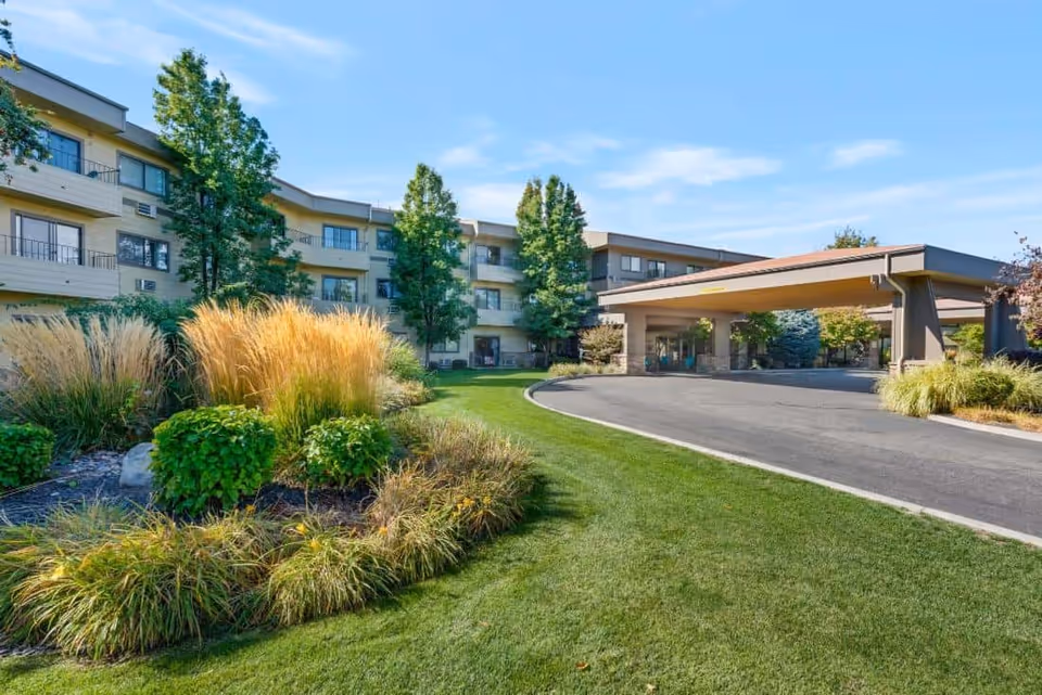 Exterior view of a senior living facility with a curved driveway, well-maintained green lawn, landscaped bushes and tall grasses, and a three-story building with balconies and large windows under a clear blue sky.