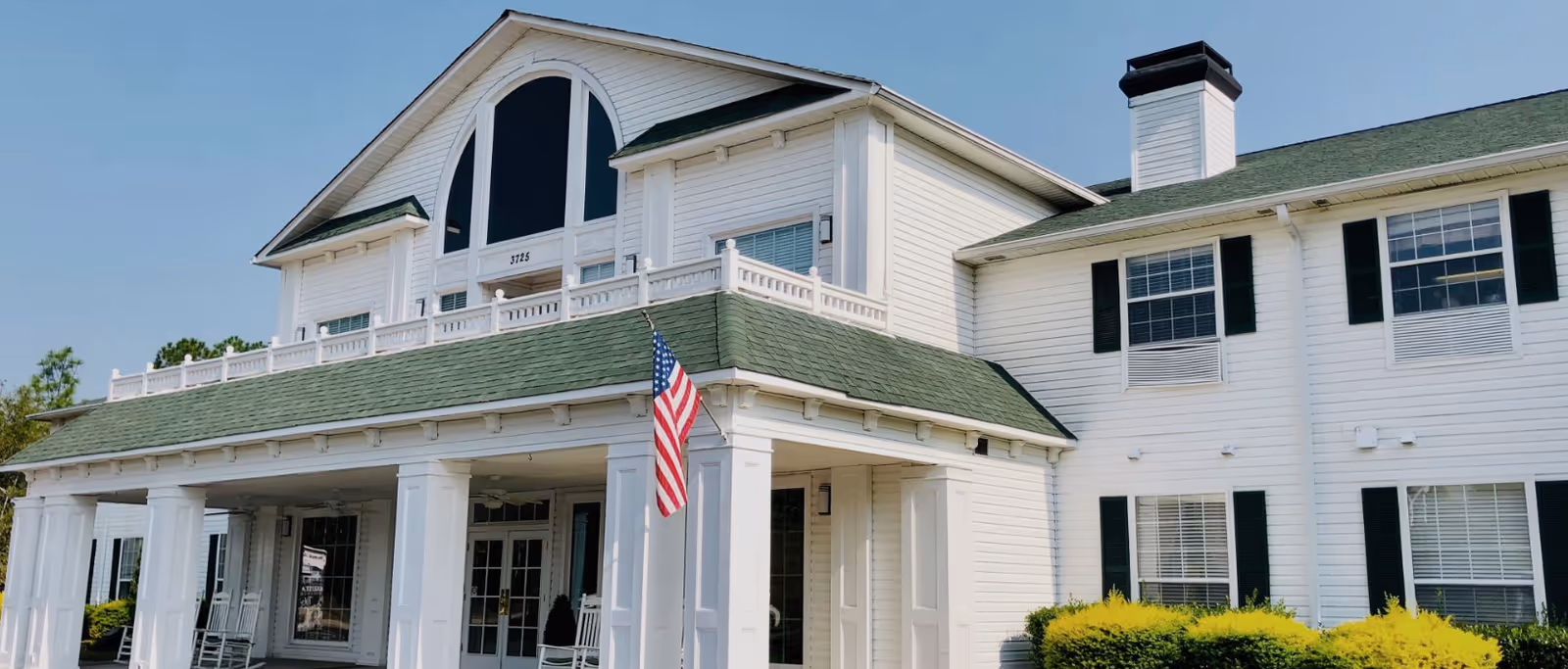 Front exterior view of a large white senior living facility building with green roof shingles, multiple windows with black shutters, a covered porch with white columns, rocking chairs, and an American flag hanging near the entrance.