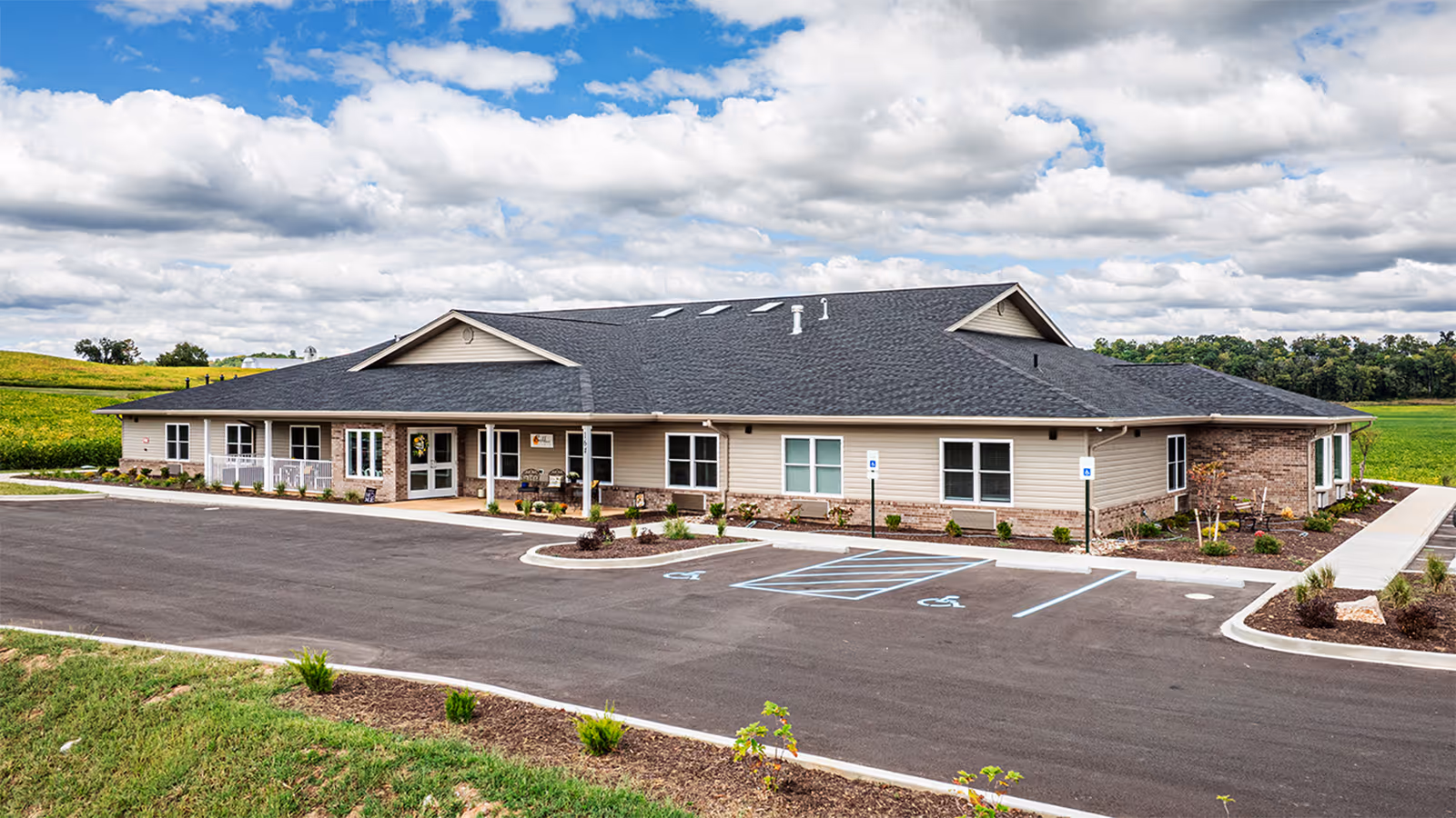 Single-story beige brick-and-siding senior living building with a paved parking lot and accessible spaces in a rural setting under a cloudy sky.