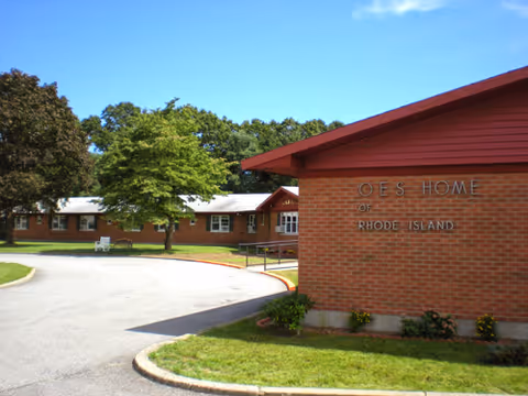 Brick senior living facility exterior with curved driveway, sign, and surrounding trees.