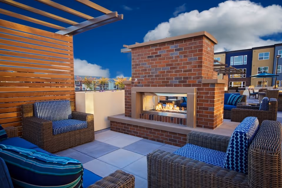 Rooftop outdoor seating area with wicker armchairs, patterned cushions, and a double-sided brick fireplace.