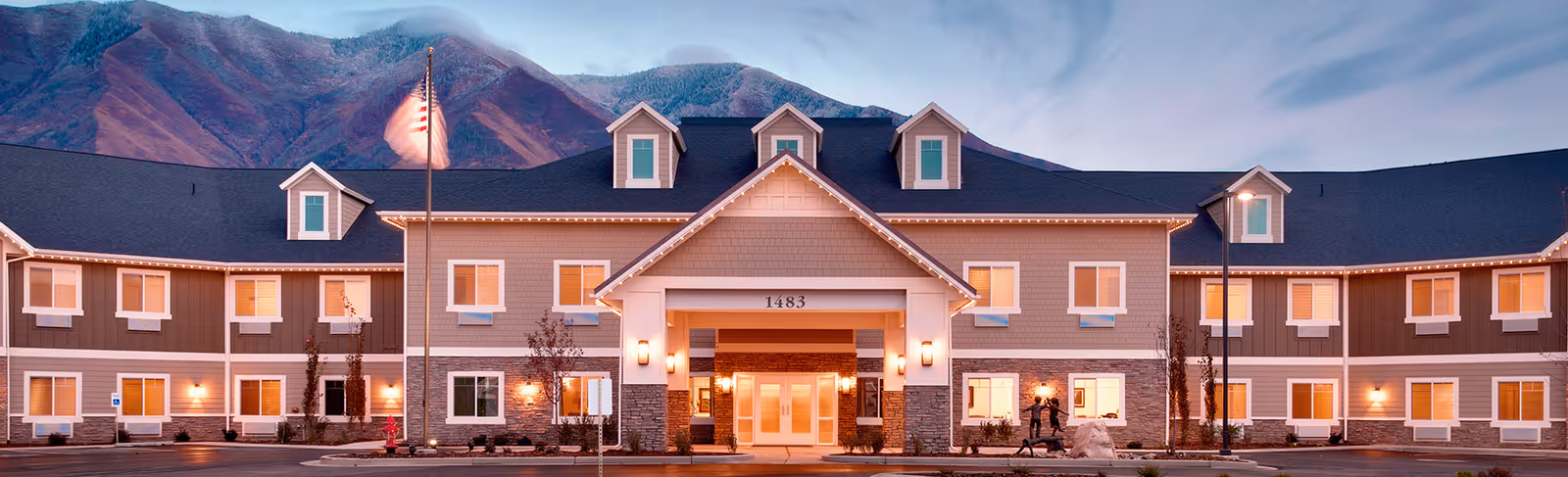Front exterior of a two-story senior living building at dusk with an illuminated covered entrance, an American flag, and mountains in the background.
