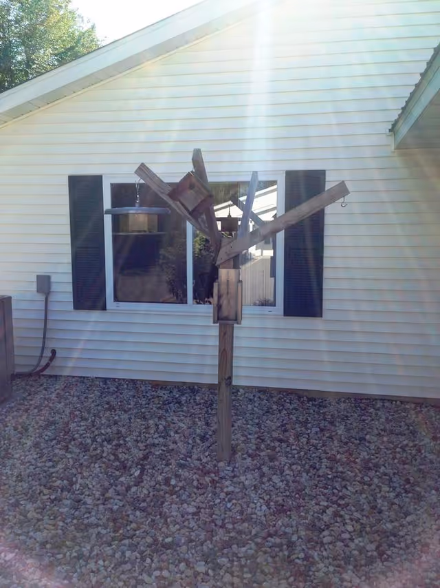 A wooden bird feeder structure with multiple feeders attached to a central post, situated on a bed of small rocks in front of a white building with black shutters and a window. Sunlight is streaming down from above, creating a bright glare.
